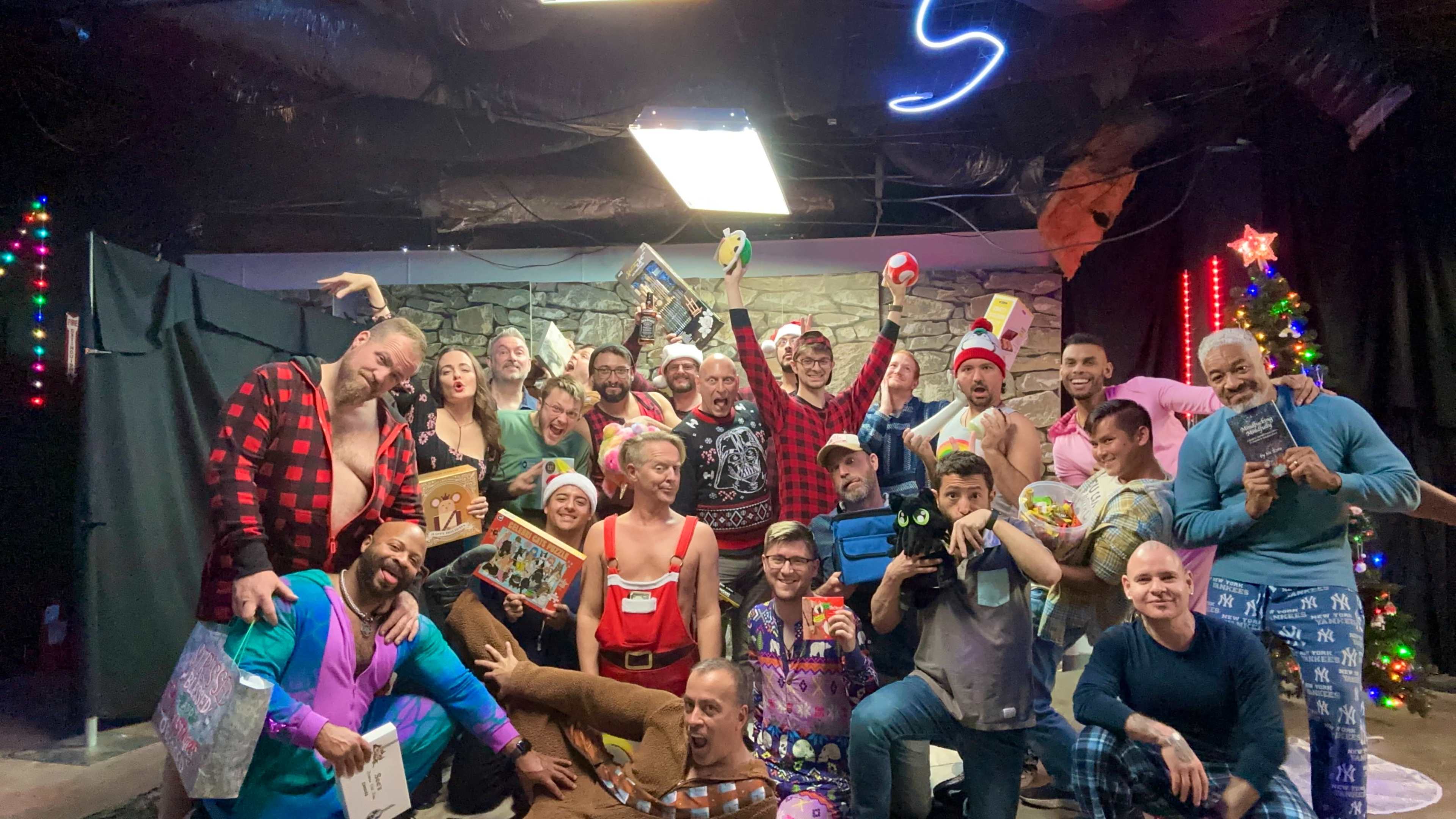 A group of people wearing festive attire and holding gifts poses together in a decorated room with holiday lights and a Christmas tree.