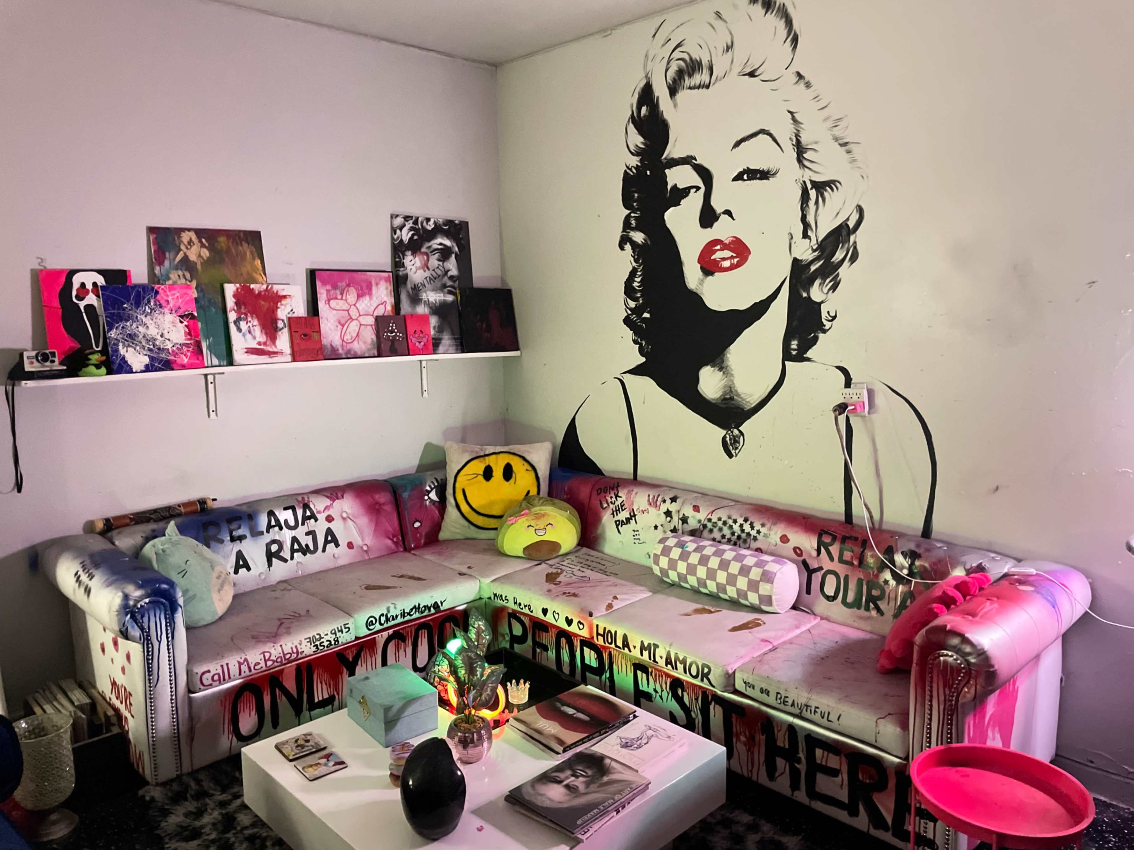 The image shows a colorful room with a large mural of a woman on the wall, a patterned couch, and various artworks displayed on shelves.