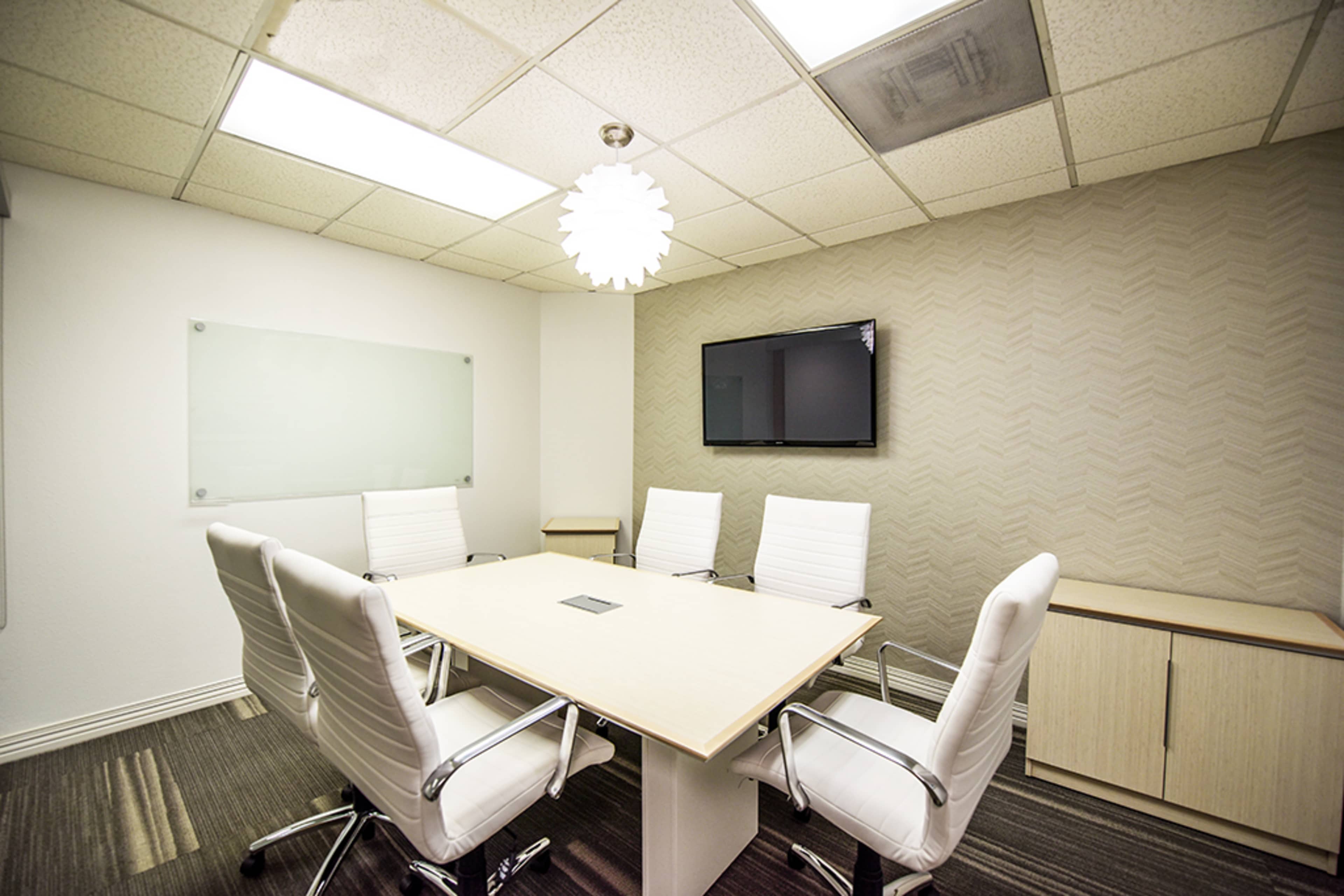 A modern conference room with a large table surrounded by white chairs, a wall-mounted television, and a decorative light fixture.