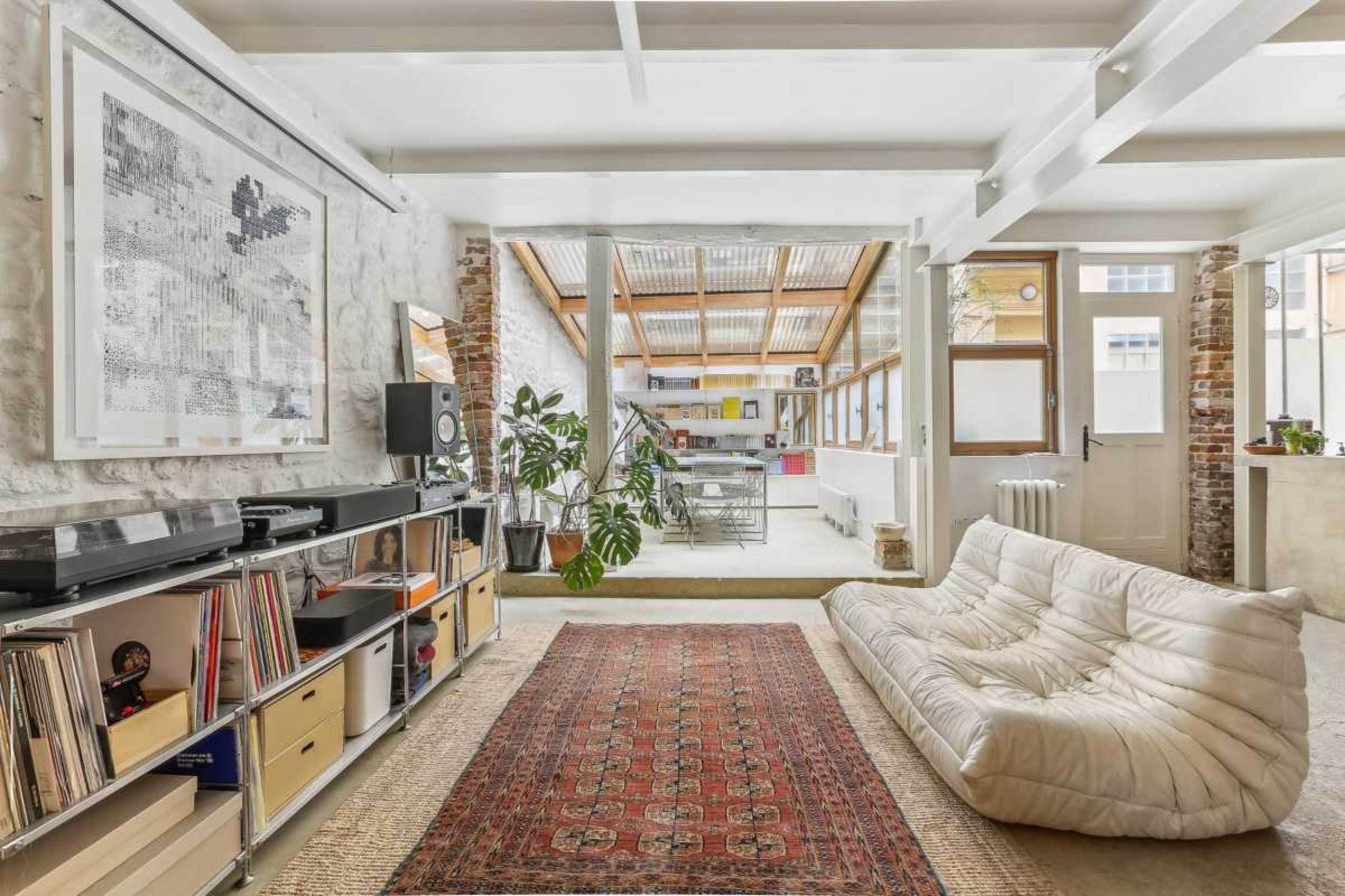 The living space features a white, minimalist couch, a record shelf with vinyl records, and a view of a sunlit room with large windows and plants in the background.