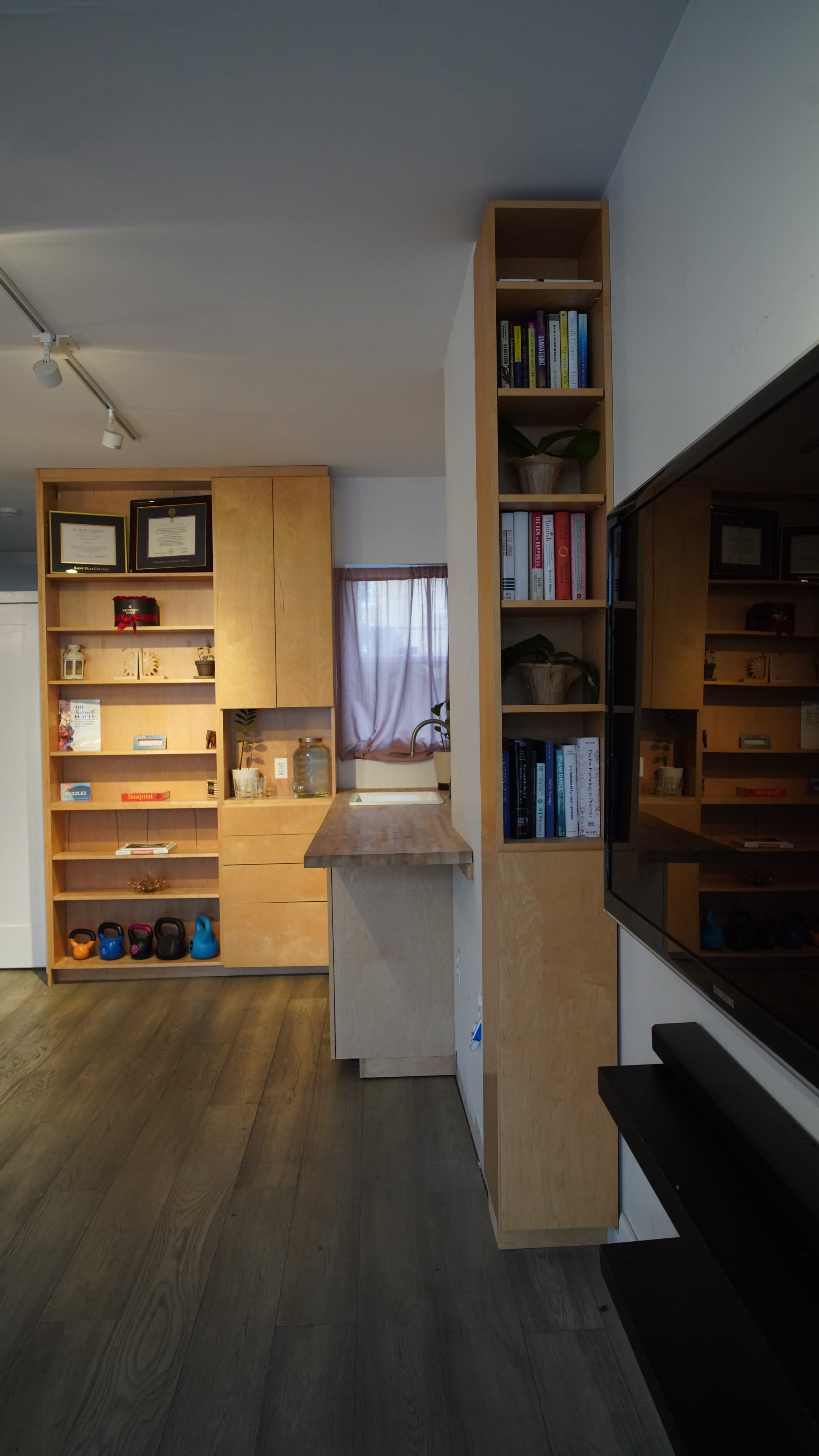 The image shows a modern interior space featuring a sleek kitchen area with wooden cabinets, a shelf filled with books and decorative items, and a large television mounted on the wall.