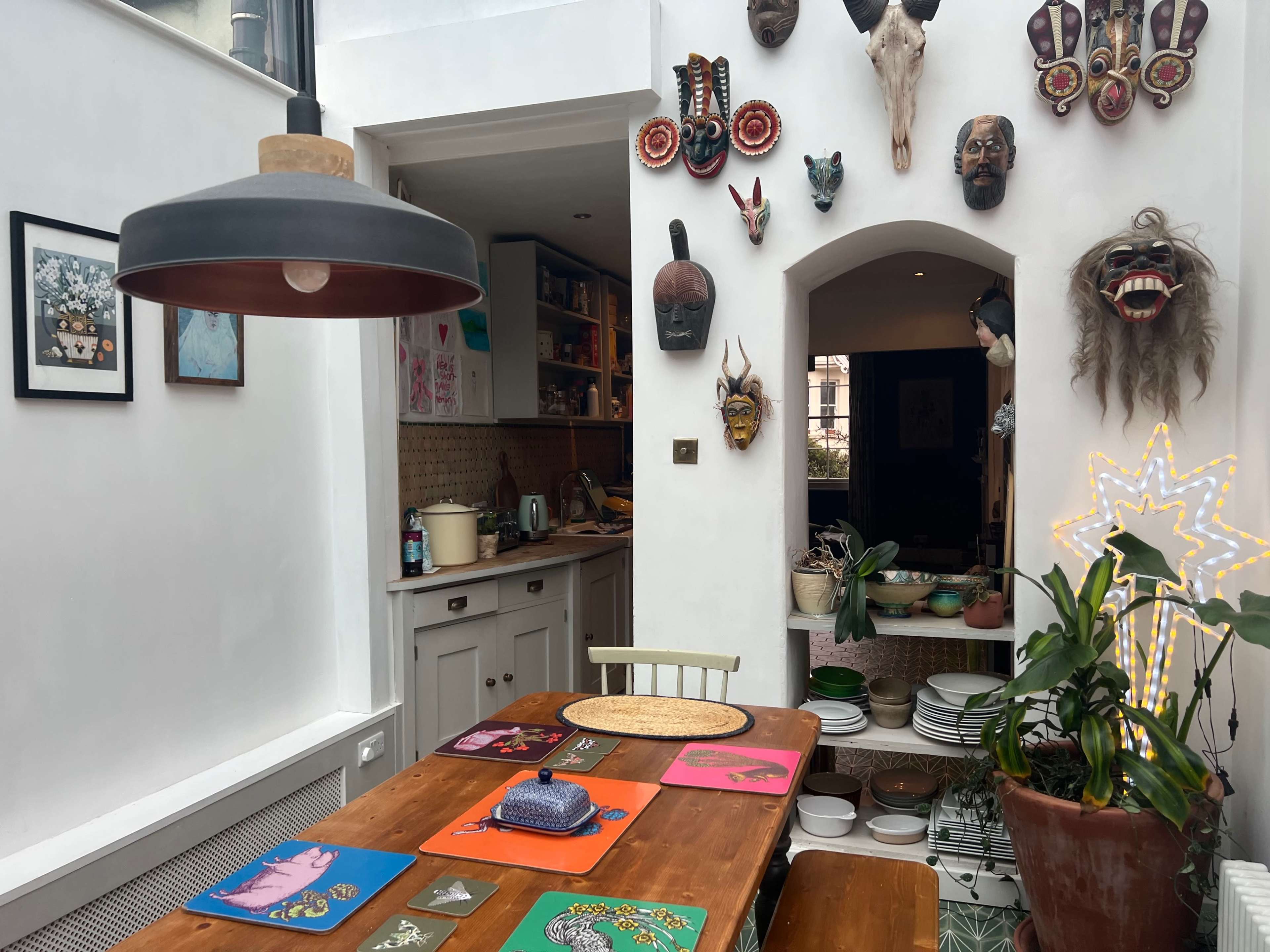 A wooden dining table is set in a brightly lit room adorned with various masks and artwork, with a kitchen visible in the background.