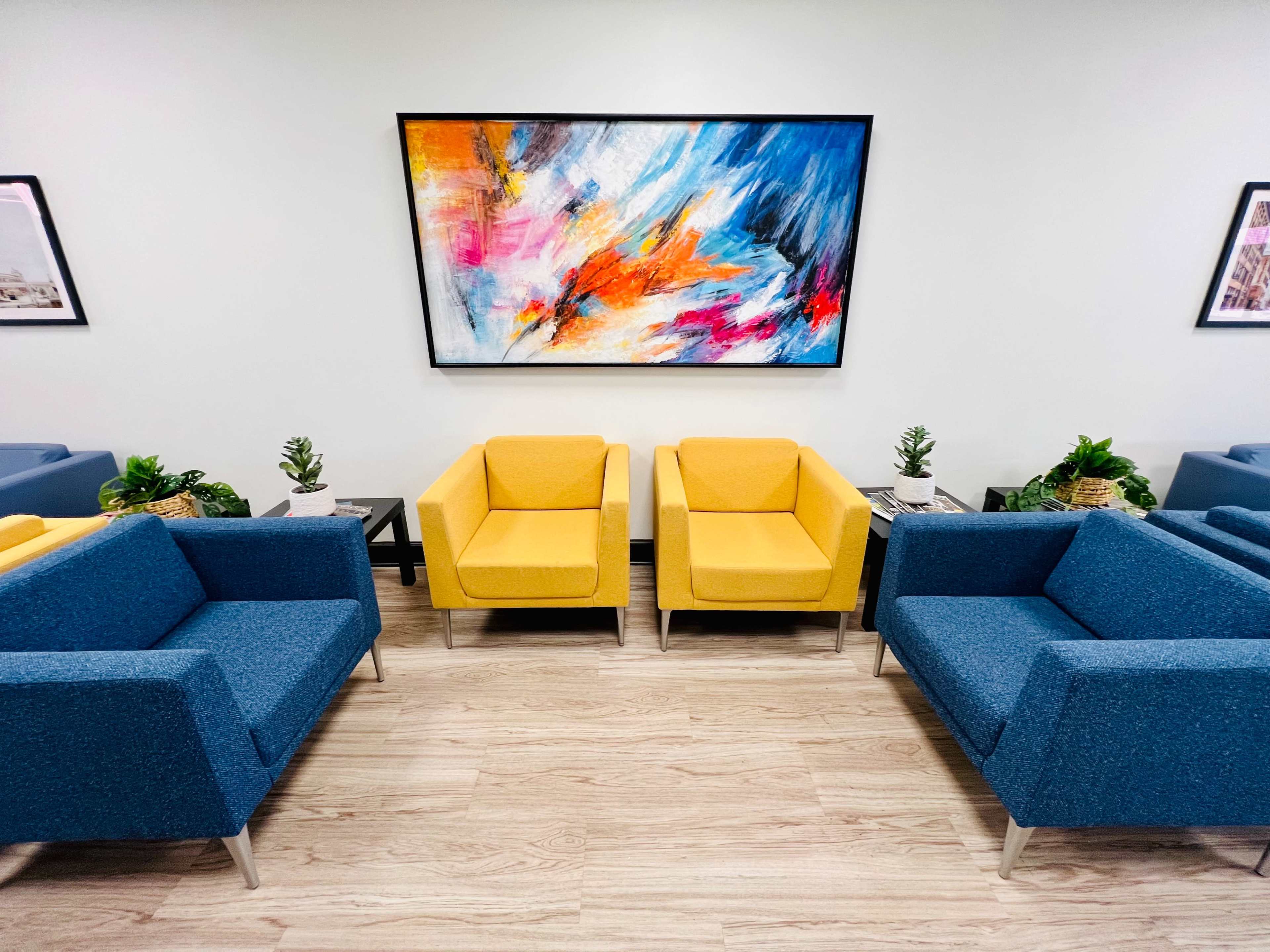 A modern waiting area with blue and yellow chairs arranged around a central table, framed by colorful artwork on the wall and potted plants.