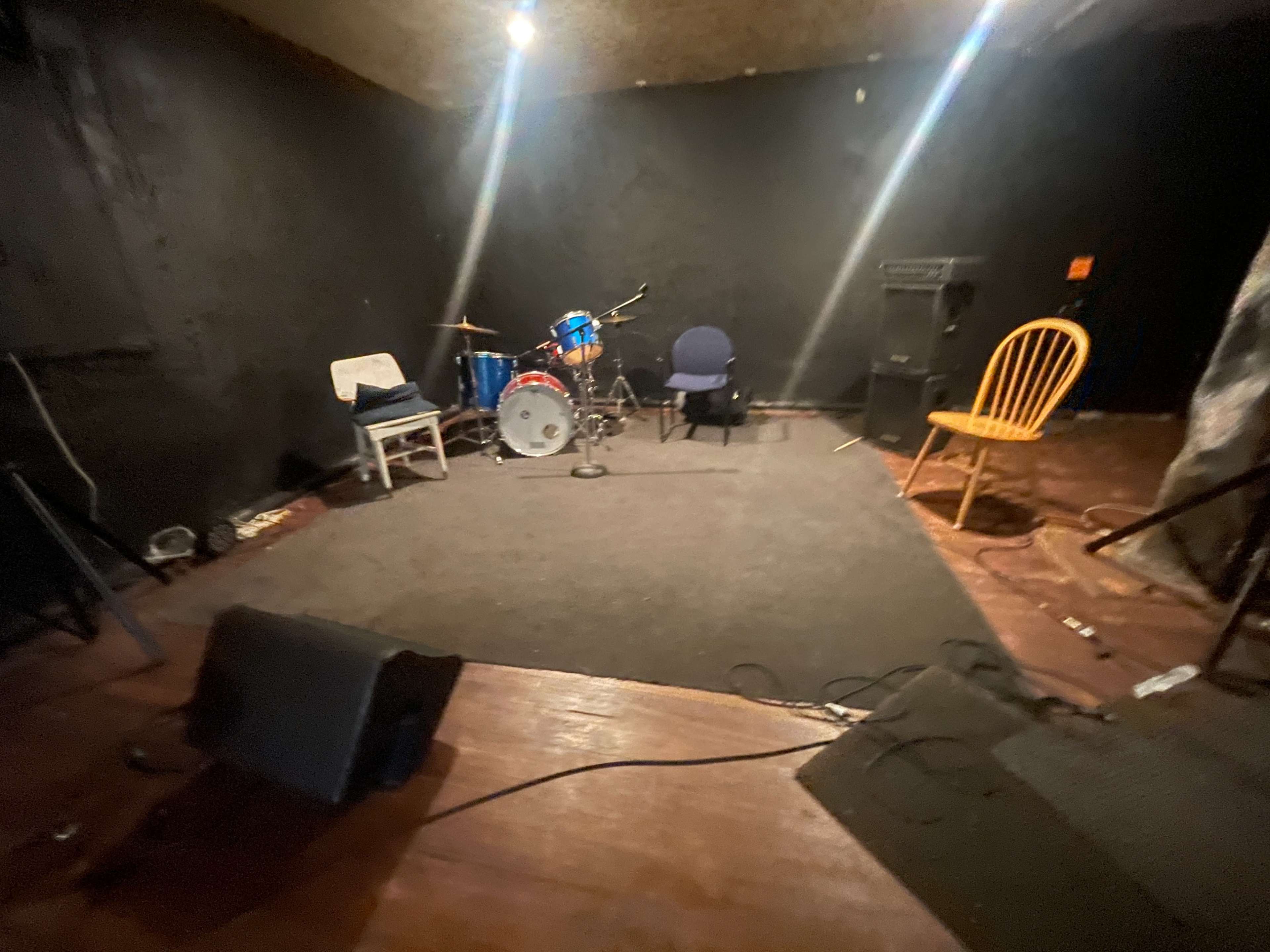 The image shows a small, dimly lit music performance space with a drum set, two chairs, and sound equipment against a black wall.