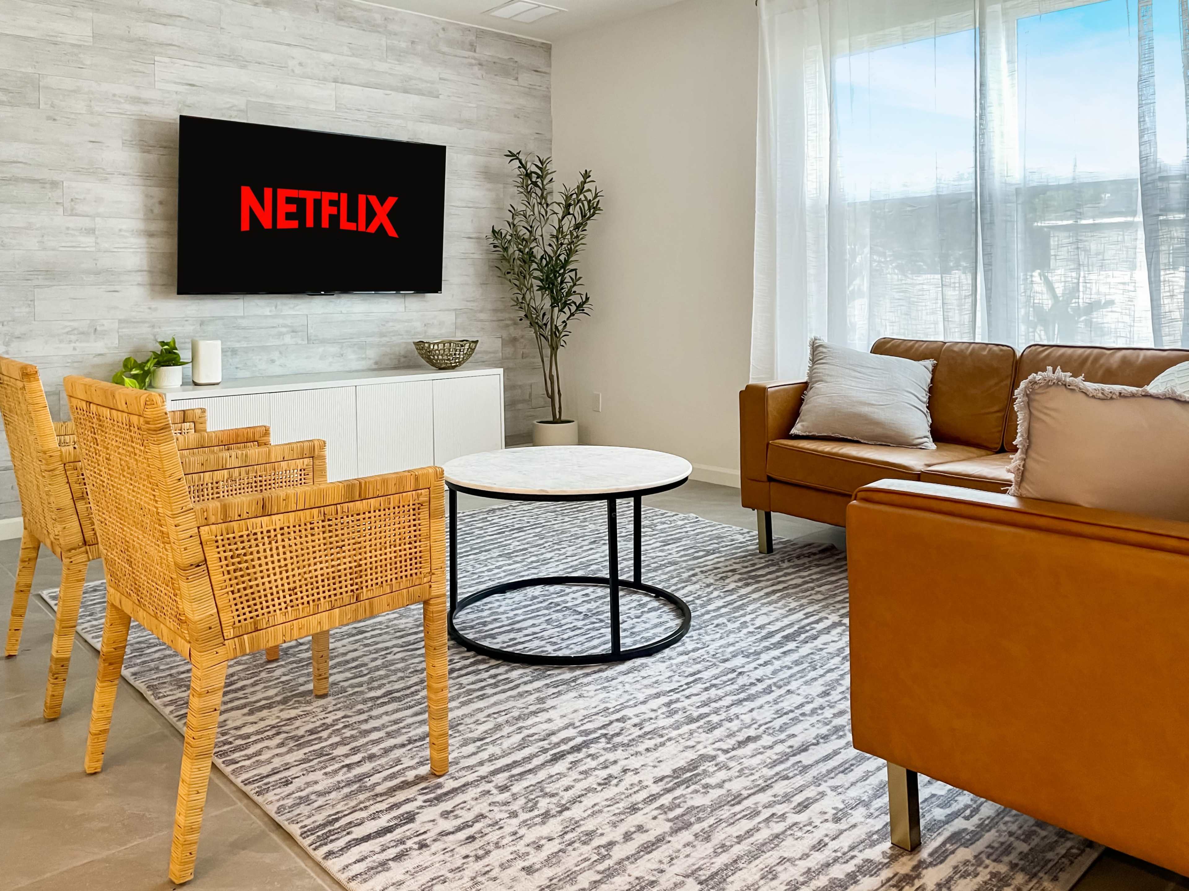 A modern living room features a television displaying the Netflix logo, a circular coffee table, and wicker chairs arranged around a light-colored rug.