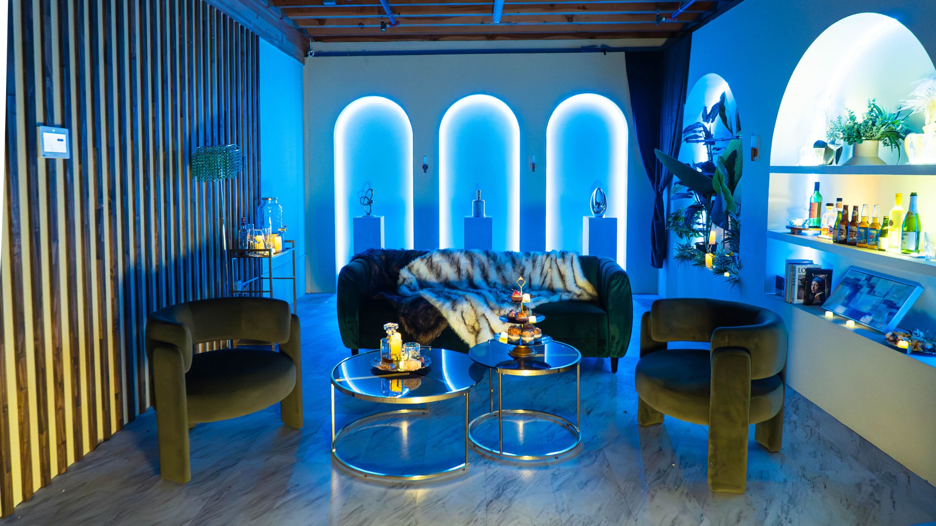 The image depicts a modern lounge area featuring a green velvet sofa, two round glass coffee tables, and stylish shelves illuminated with LED lighting, all set against a backdrop of wooden accents and blue tones.