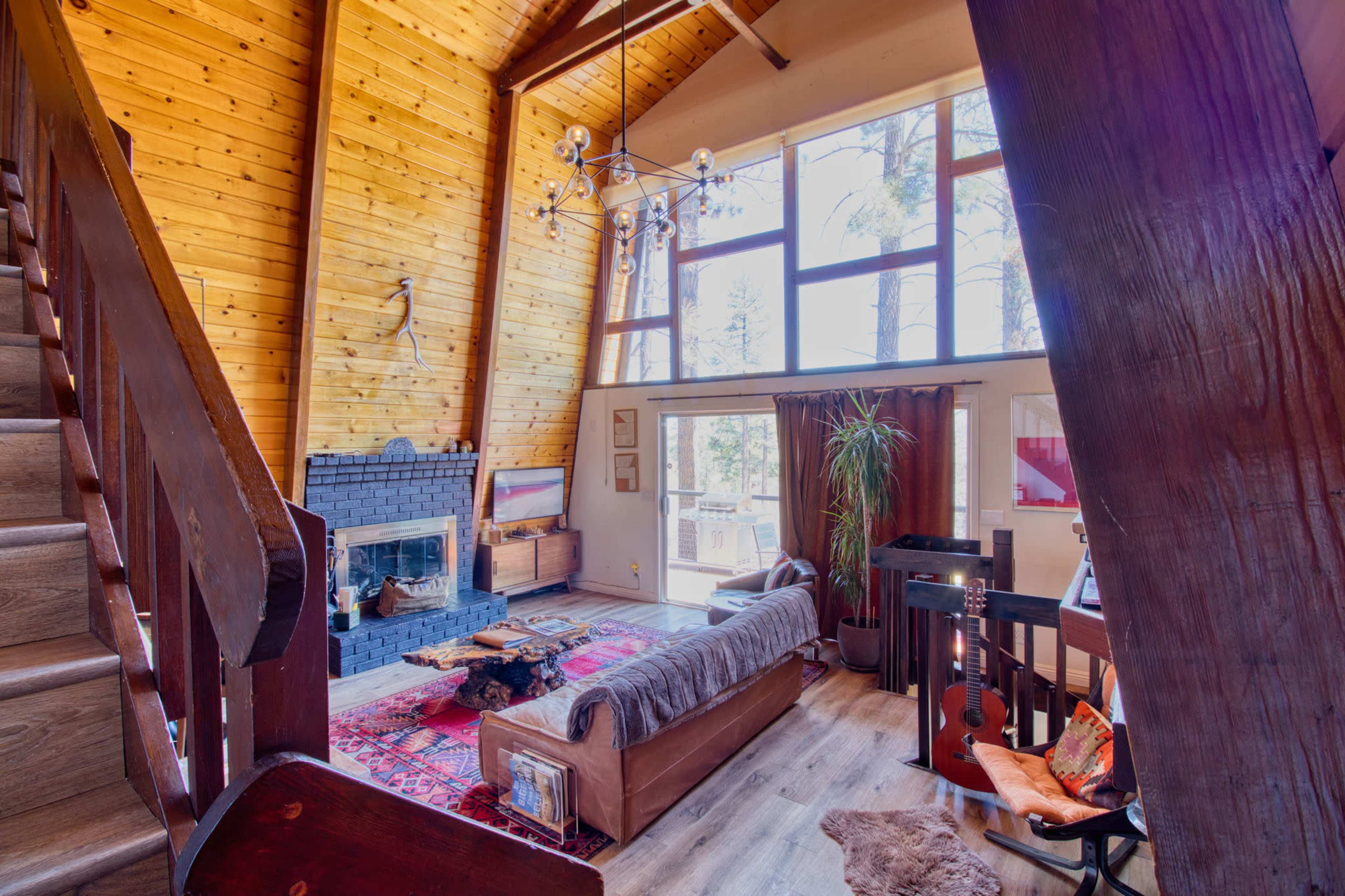 The image shows a cozy living room with a stone fireplace, wooden beams, a staircase, and large windows that let in natural light.