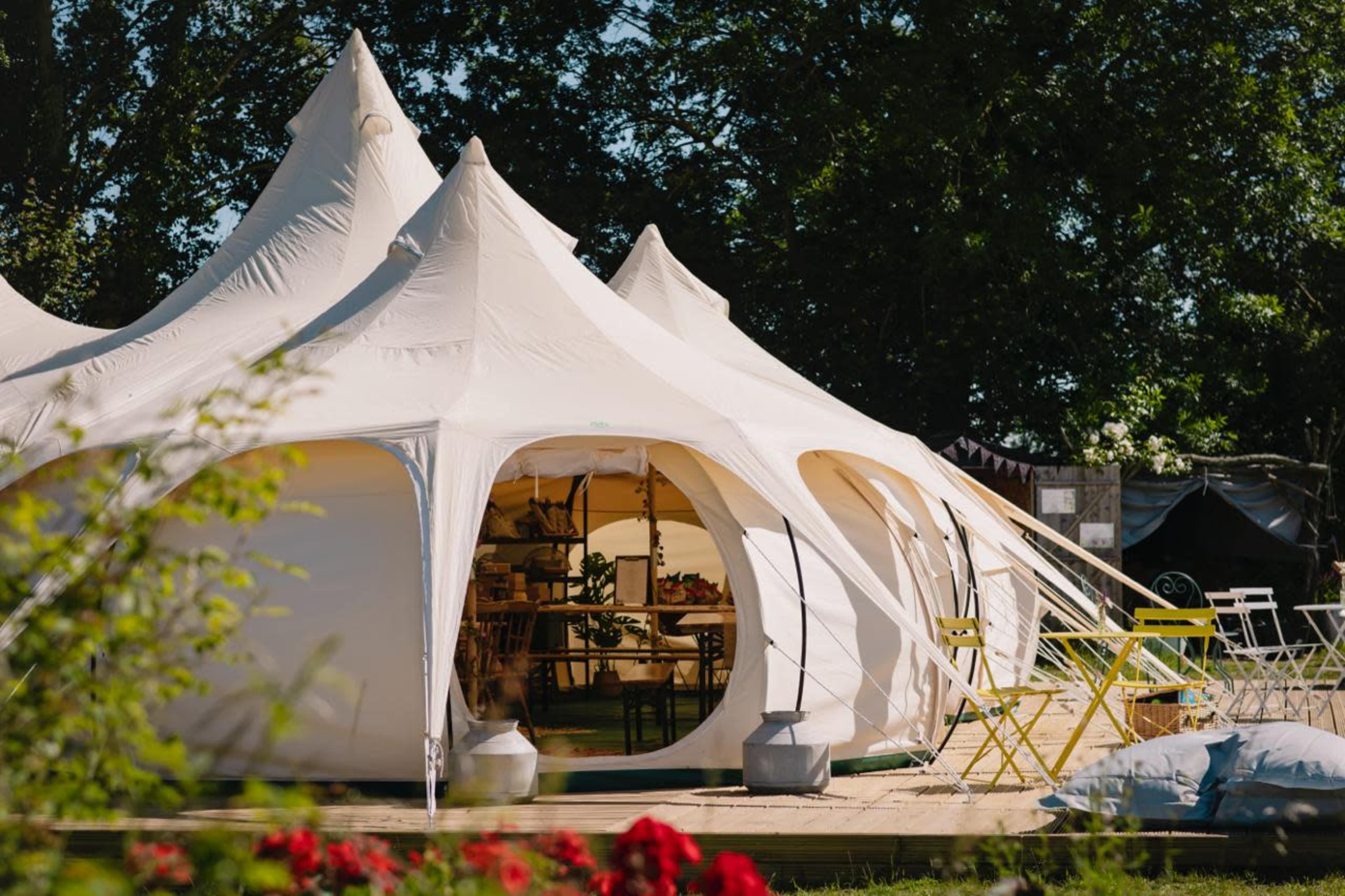 A large, white marquee tent with pointed tops is set up in an outdoor area surrounded by greenery and colorful furniture.