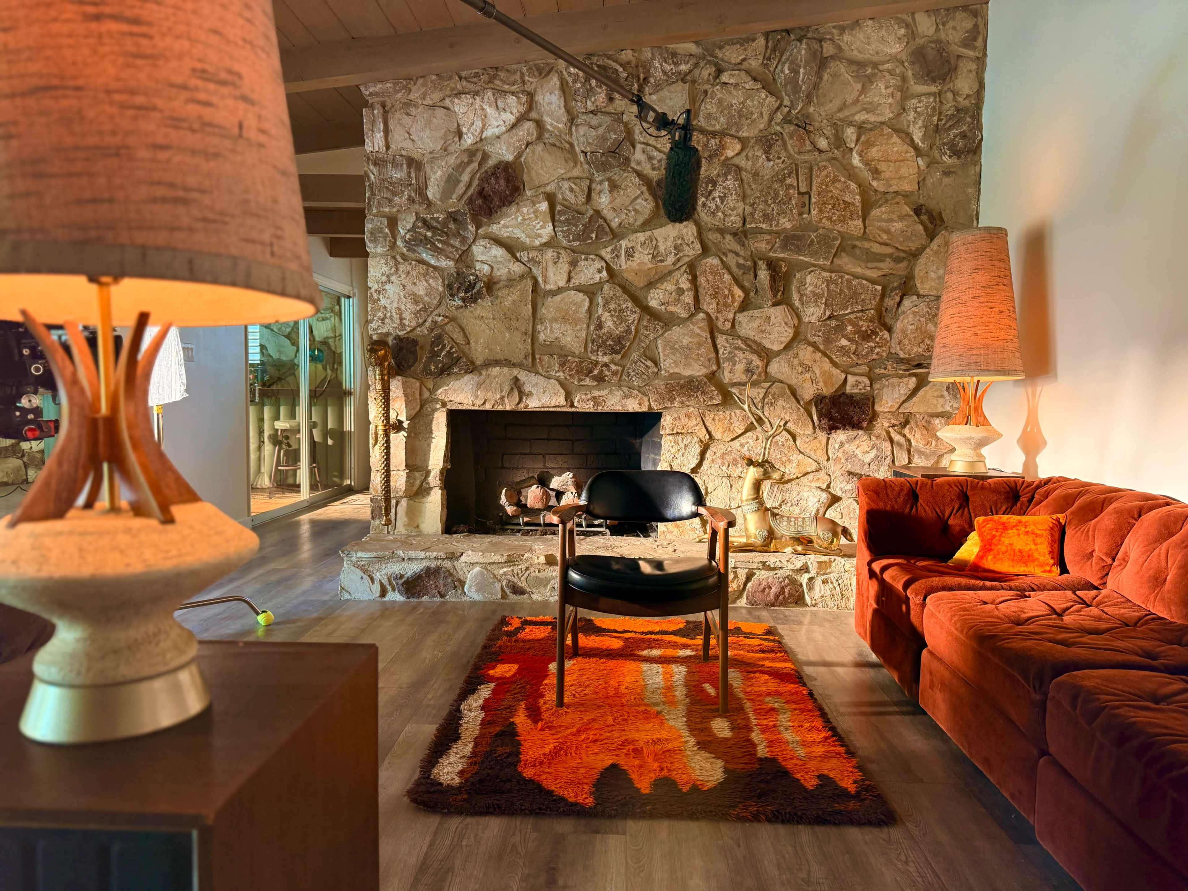 The image shows a cozy living room featuring a stone fireplace, an orange rug, a black chair in the center, and a brown couch with matching lamps.