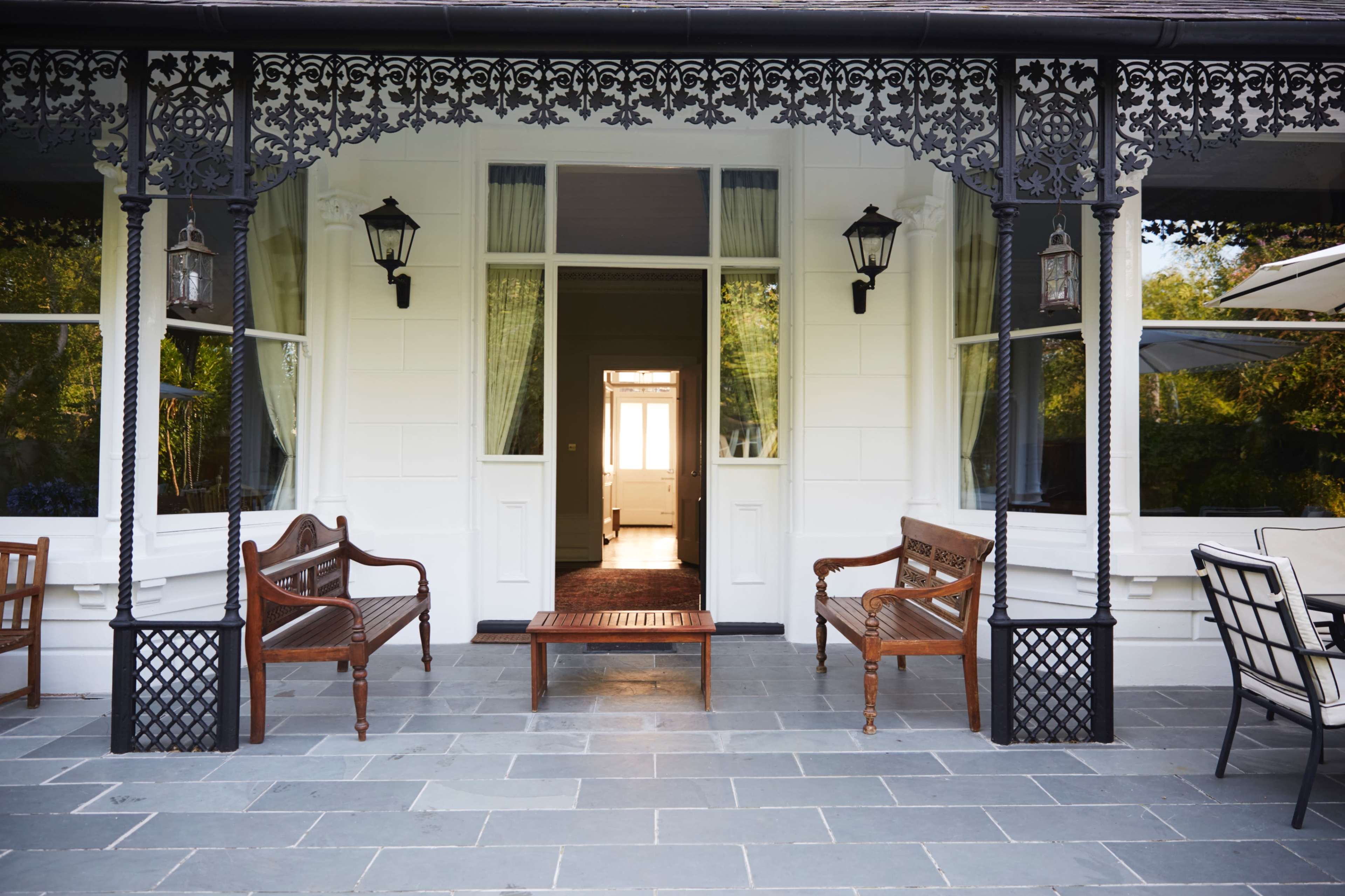A porch with two wooden chairs and a small table in front of a white house with decorative black wrought iron accents and large windows.