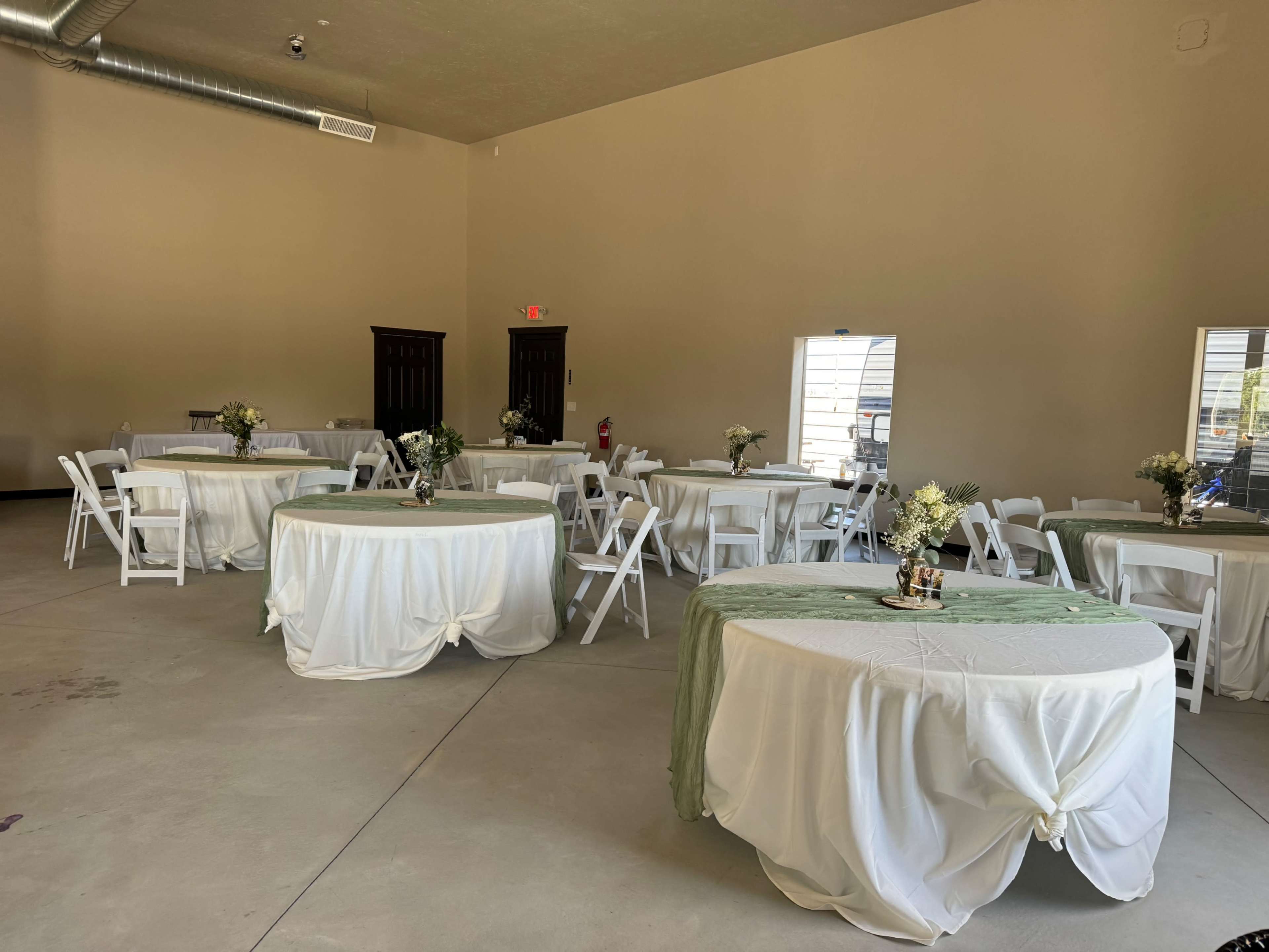 The image shows a spacious event room with several round tables covered with white tablecloths and centerpieces featuring flowers.