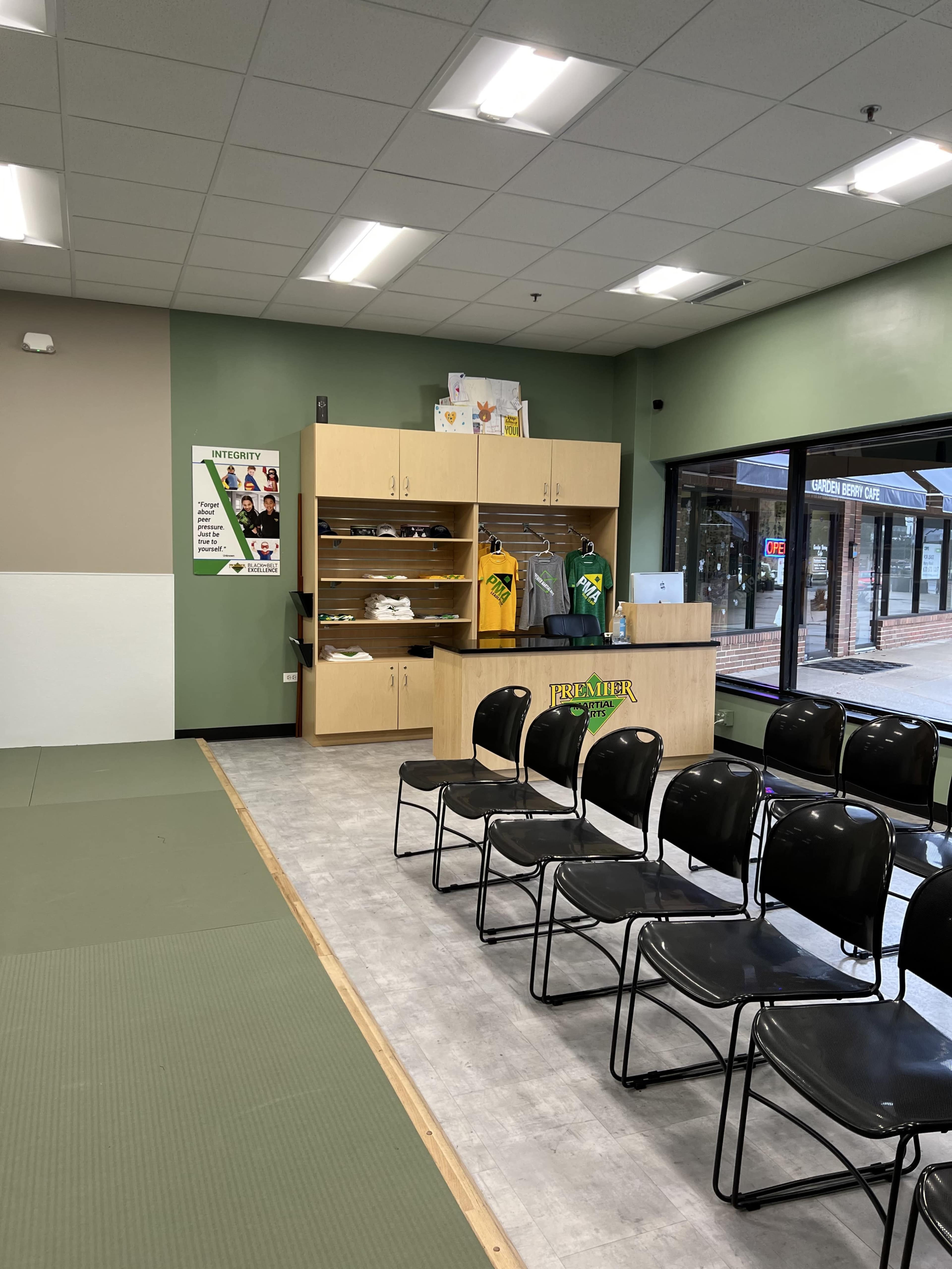 The image shows a brightly lit dojo with a seating area lined with black chairs and a wooden reception desk displaying merchandise and promotional materials.