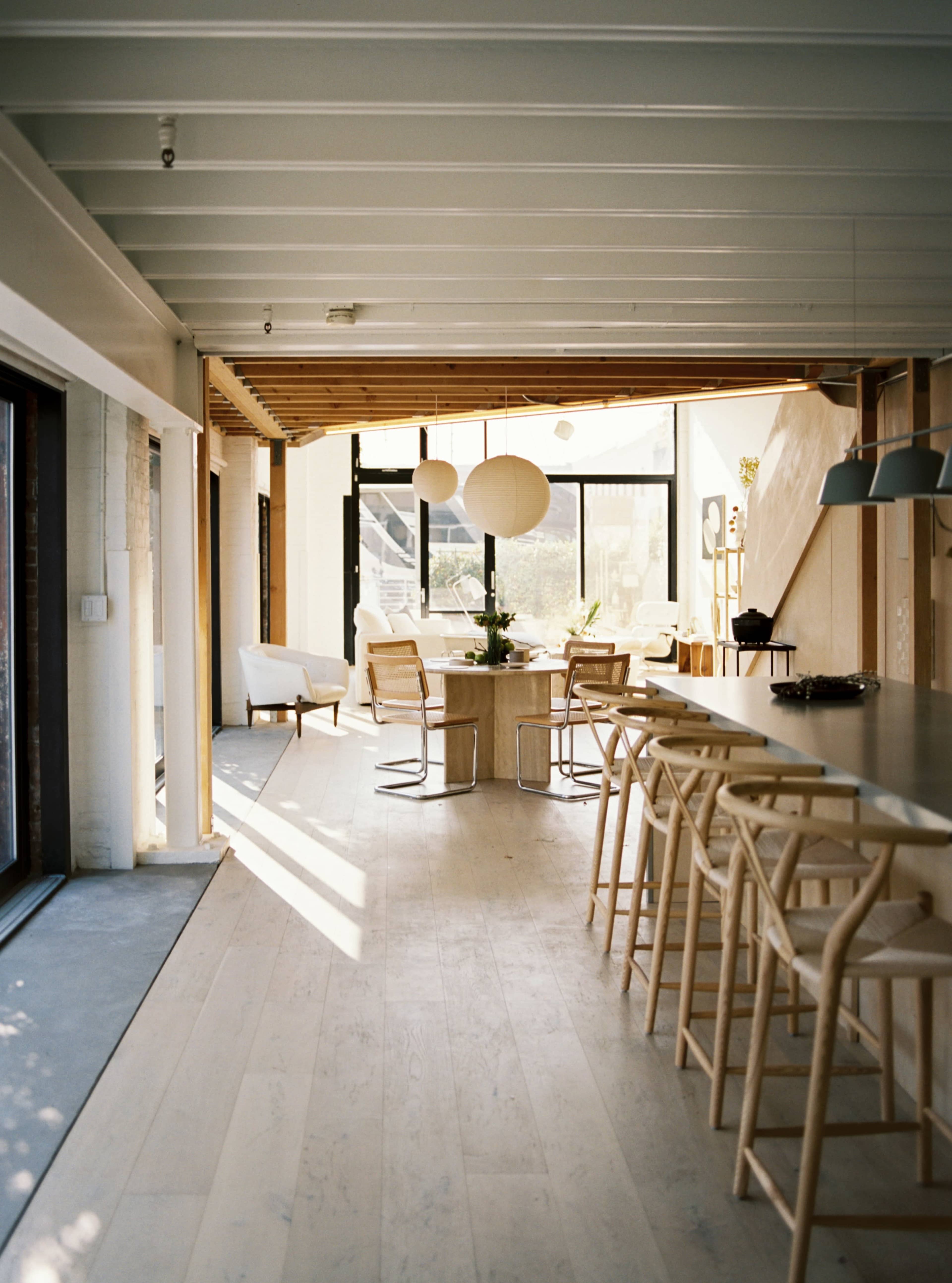 A bright, modern interior of a dining area with wooden accents, featuring a long bar with stools and a dining table surrounded by chairs, illuminated by natural light from large windows.