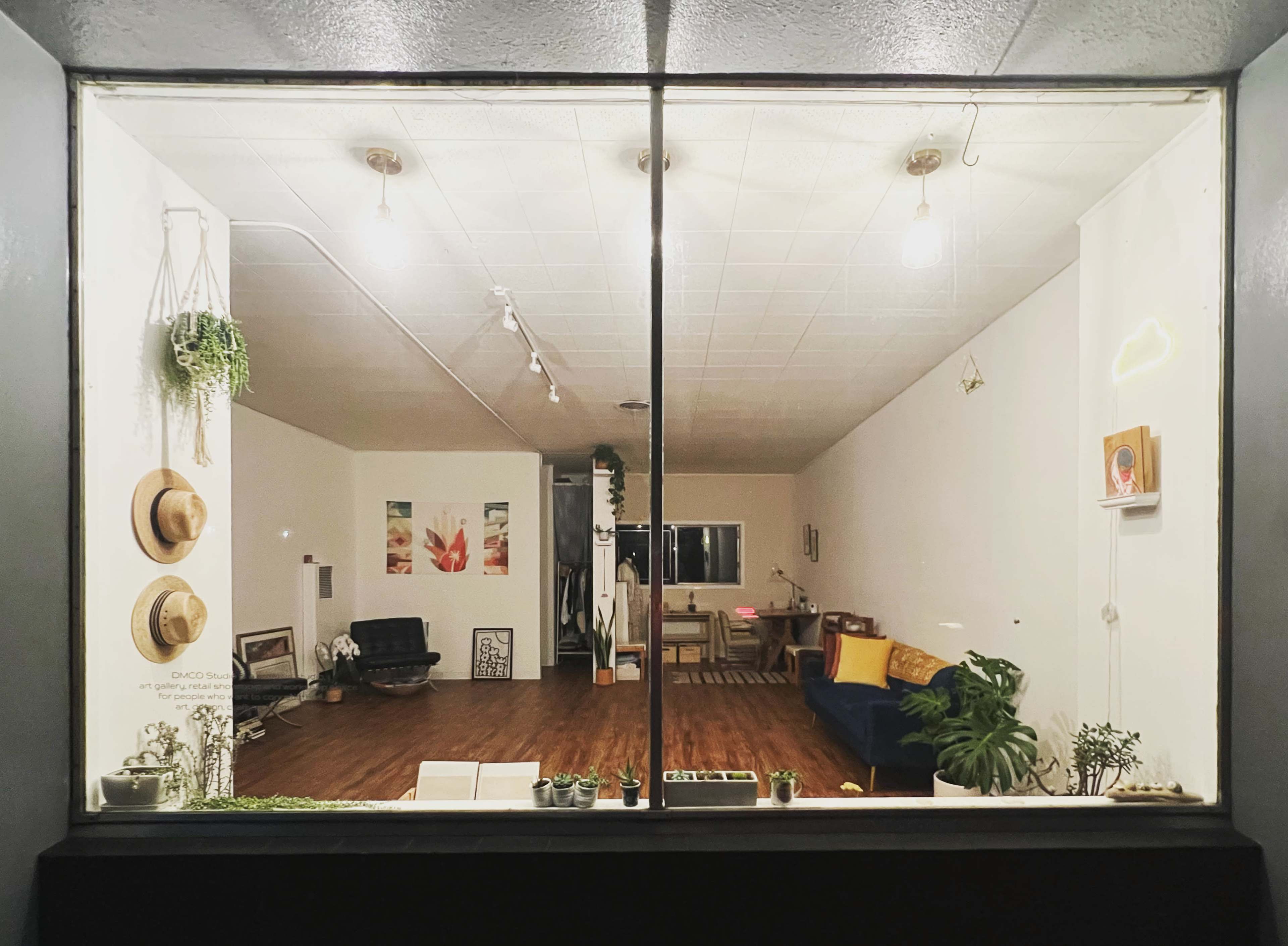 A cozy room visible through a large window, featuring a wooden floor, a blue couch, several hanging plants, and artwork on the walls.