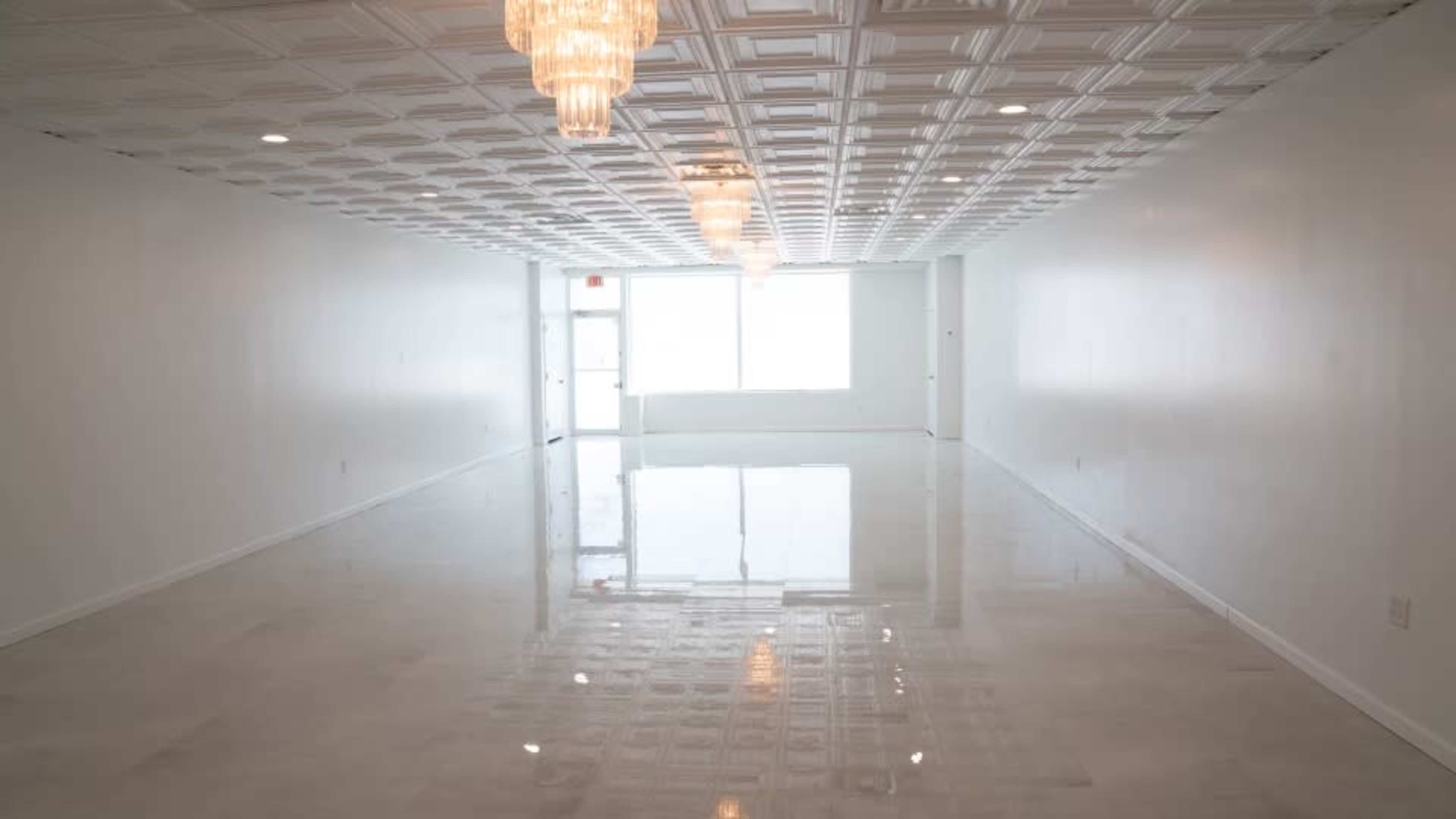 The image shows a spacious, empty room with a glossy floor, a decorative ceiling, and chandeliers hanging from above.