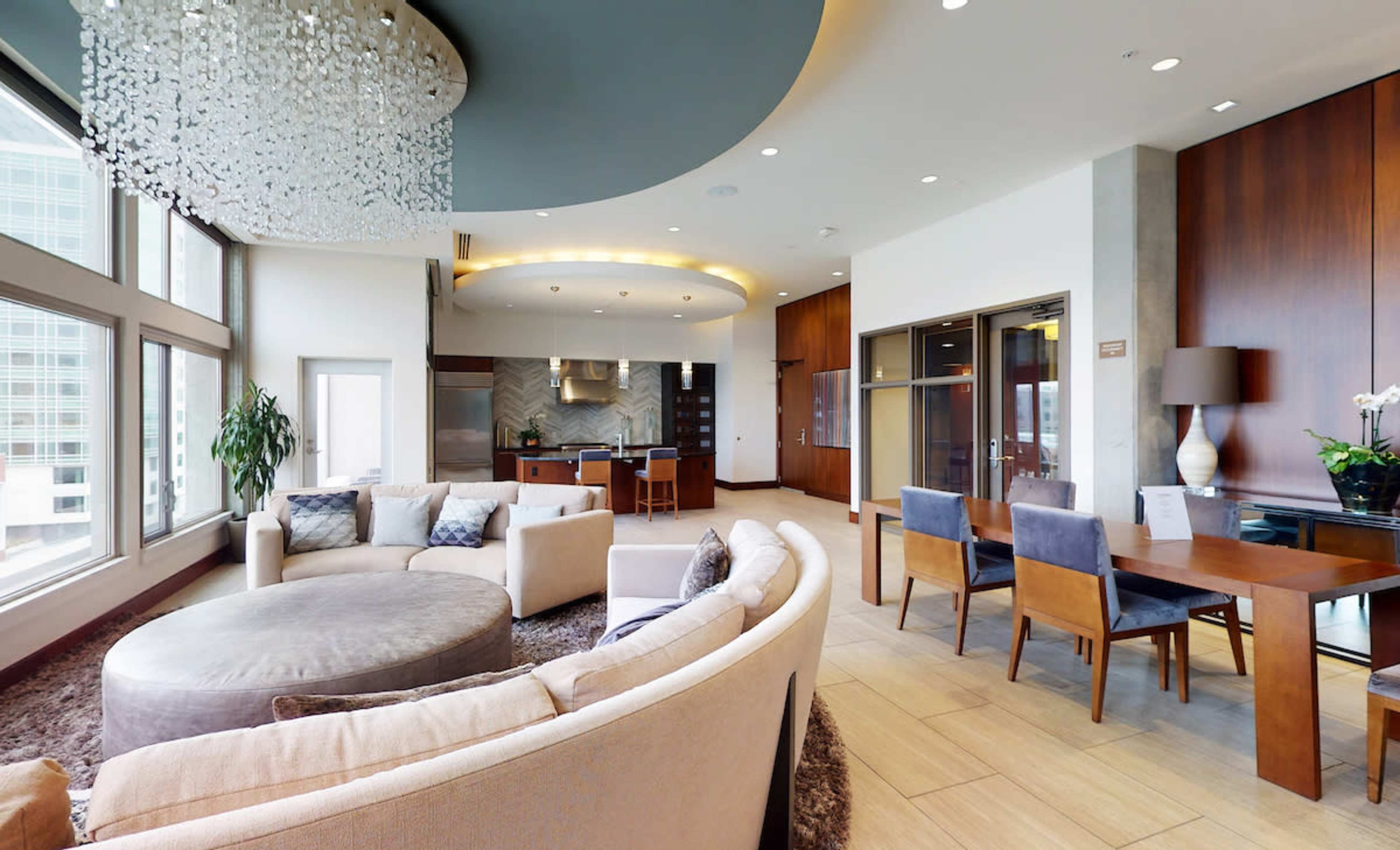 A modern, spacious living area features a large circular sofa, a dining table for four, and large windows with a view of the city.