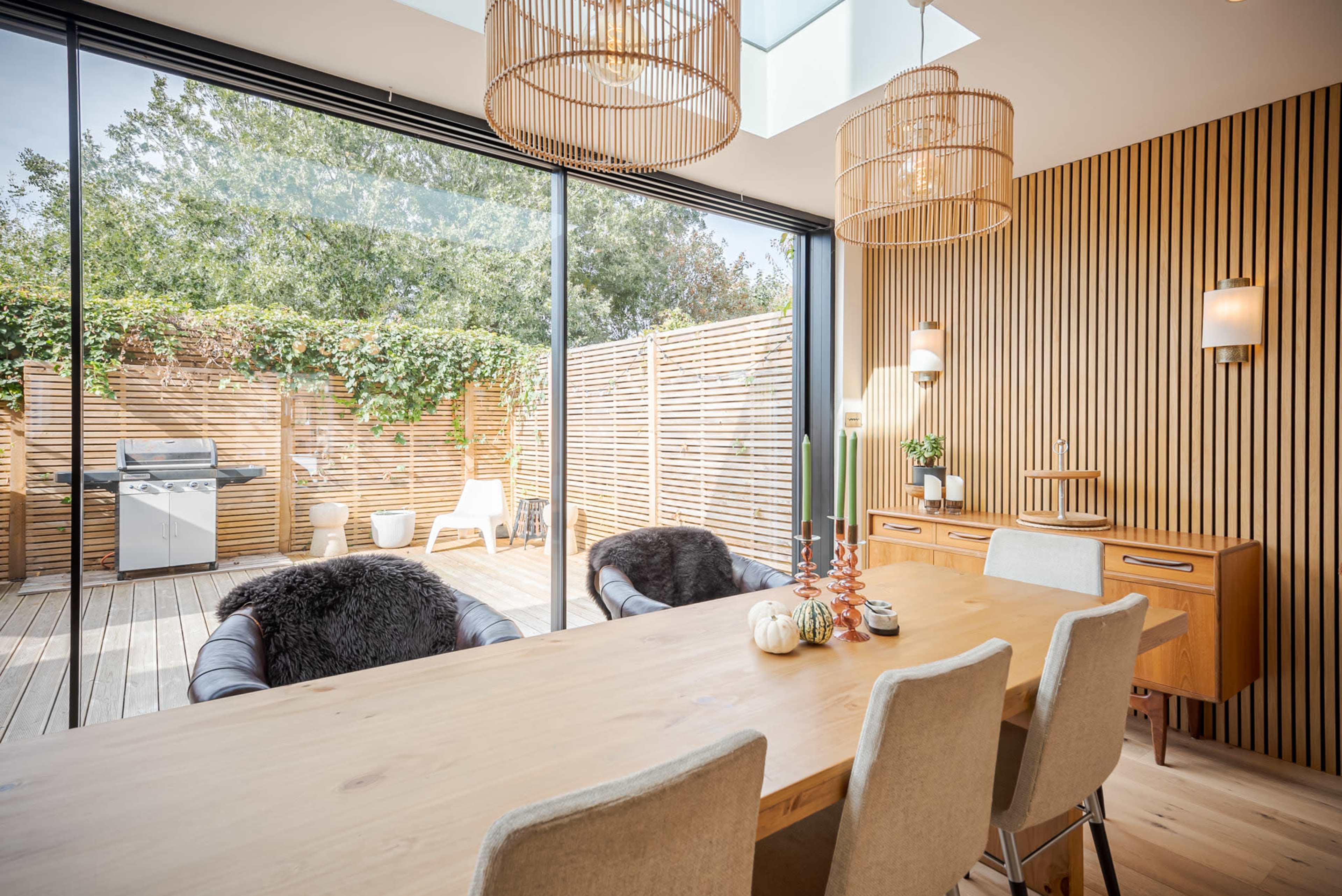 A modern dining area features a wooden table surrounded by upholstered chairs, with large glass doors opening to a patio adorned with greenery and outdoor furniture.