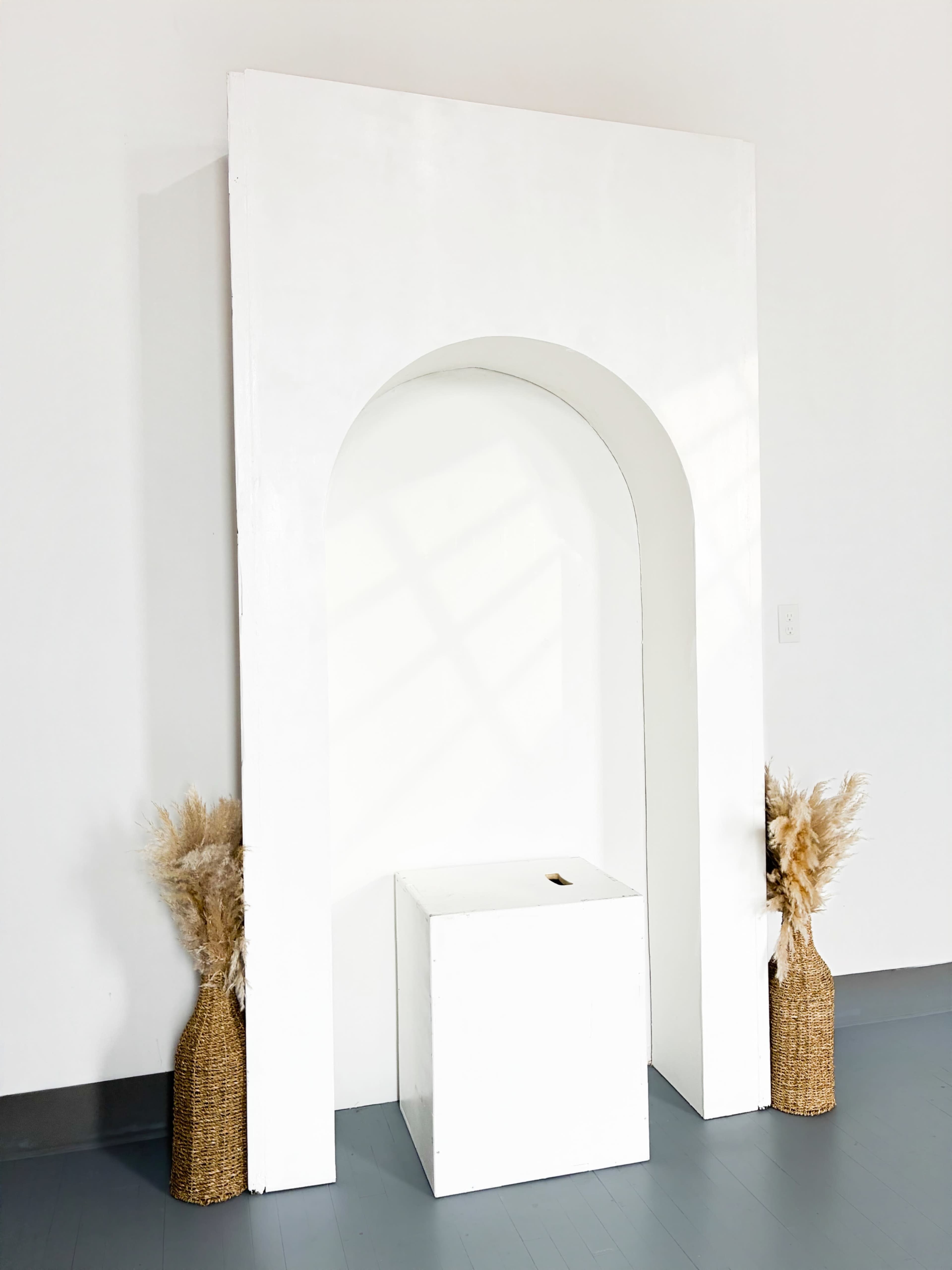 The image shows a white archway structure with a square pedestal at its base, flanked by two decorative baskets of dried pampas grass.