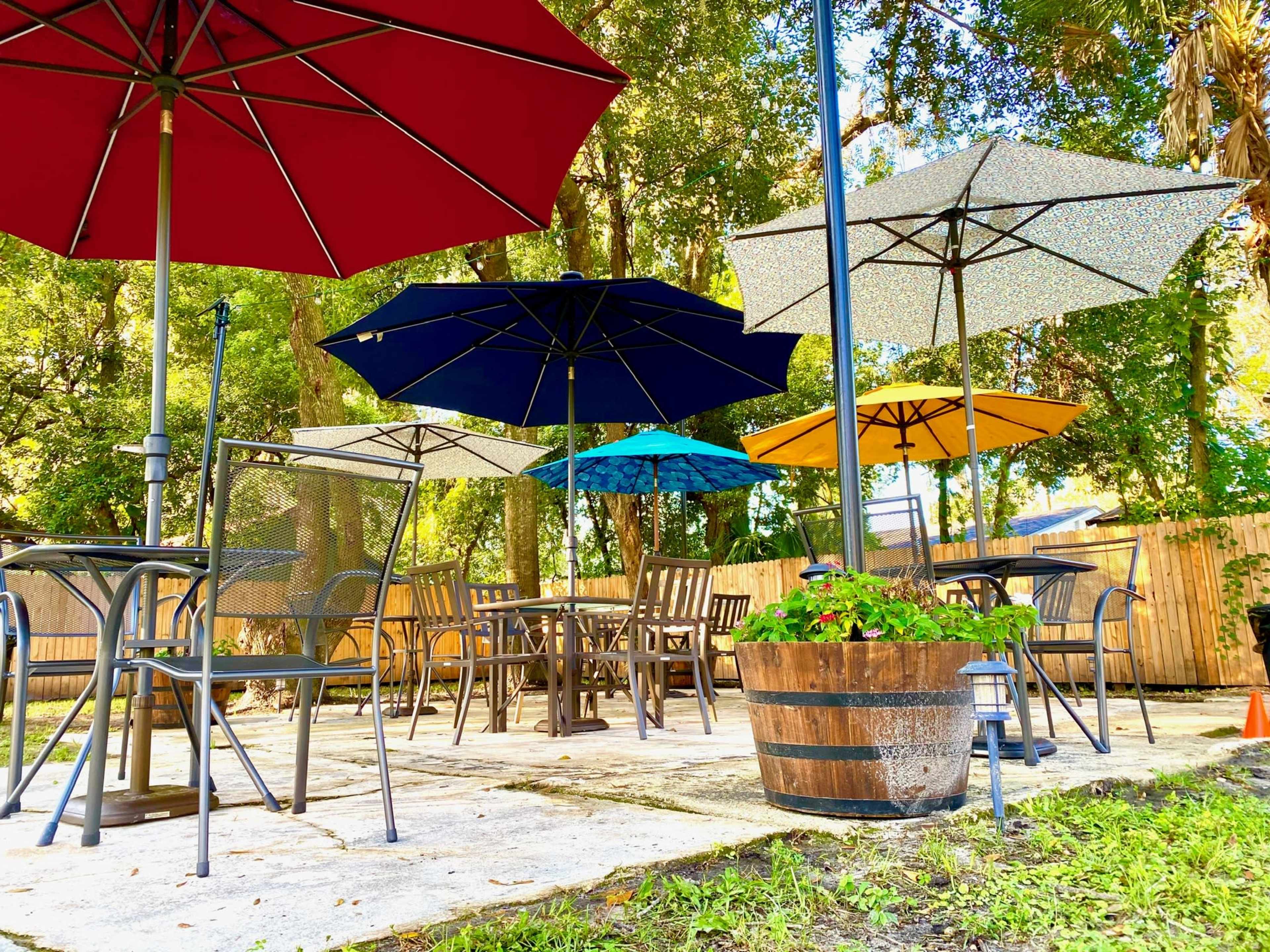A patio area features several tables with umbrellas of various colors surrounded by greenery.