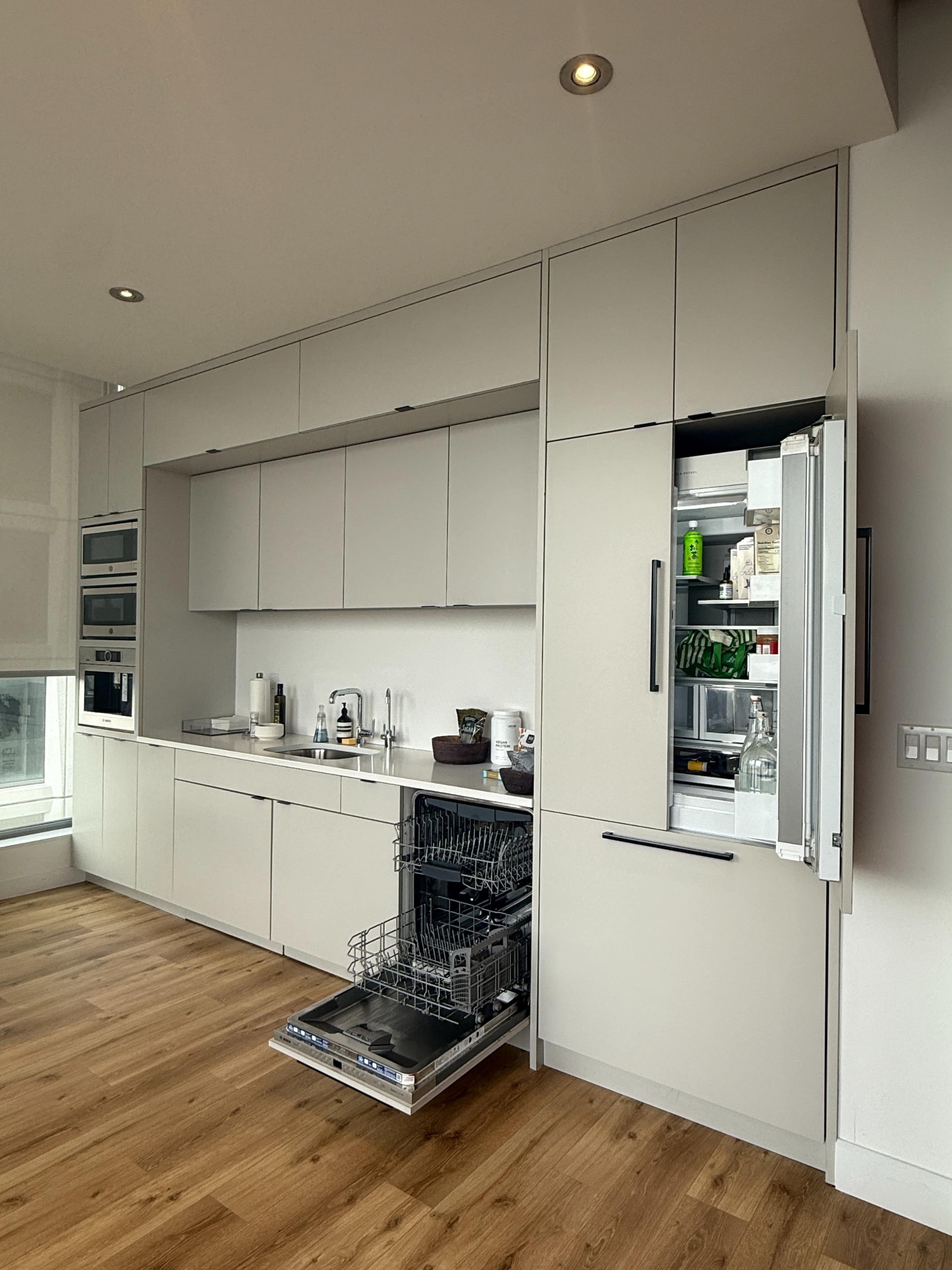 The image shows a modern kitchen with sleek cabinetry, an open dishwasher, and a refrigerator with its door ajar, set against a backdrop of wooden flooring.