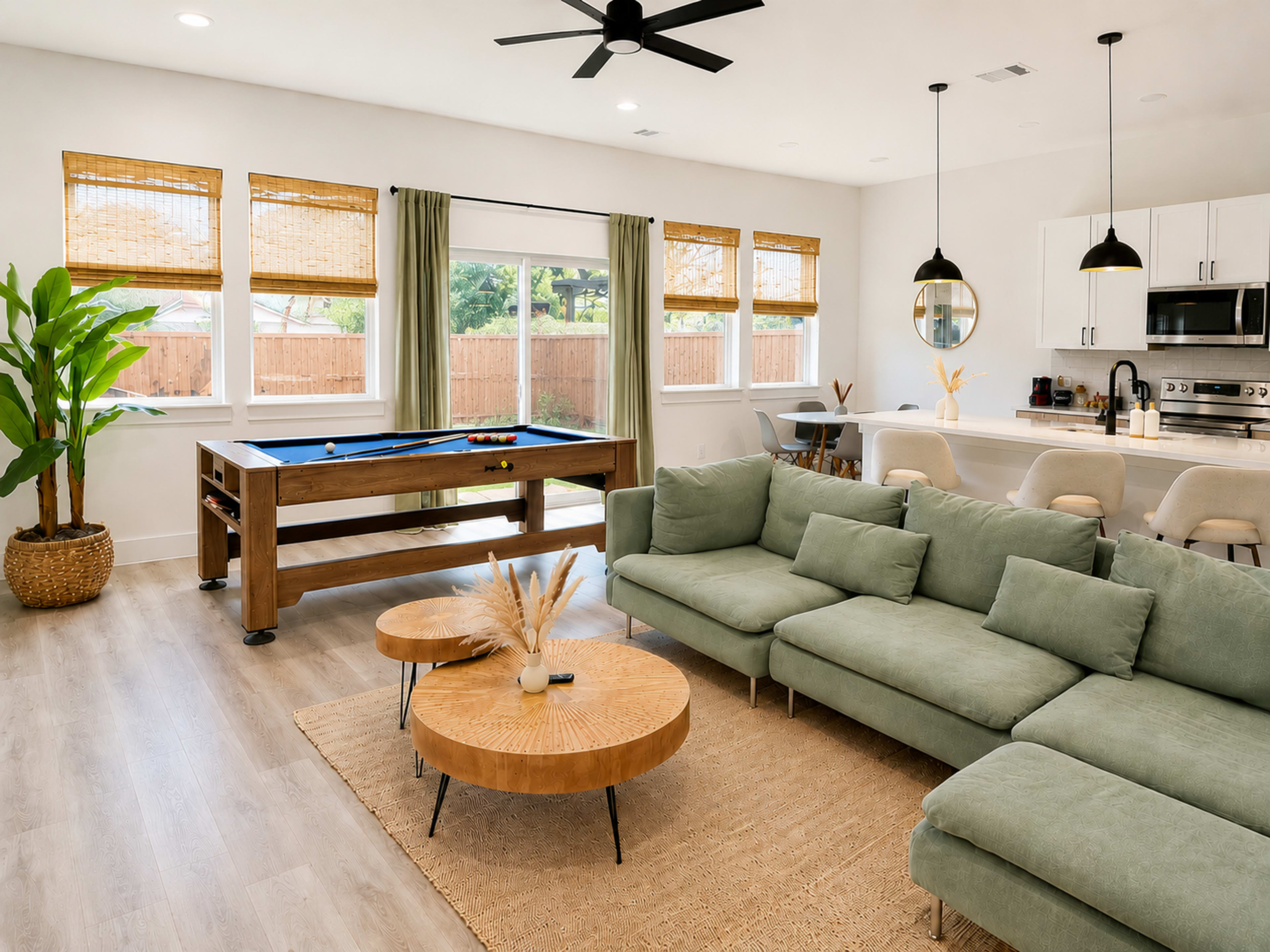 The image shows a modern living area with a sectional sofa, wooden coffee tables, a pool table, and large windows with natural light.