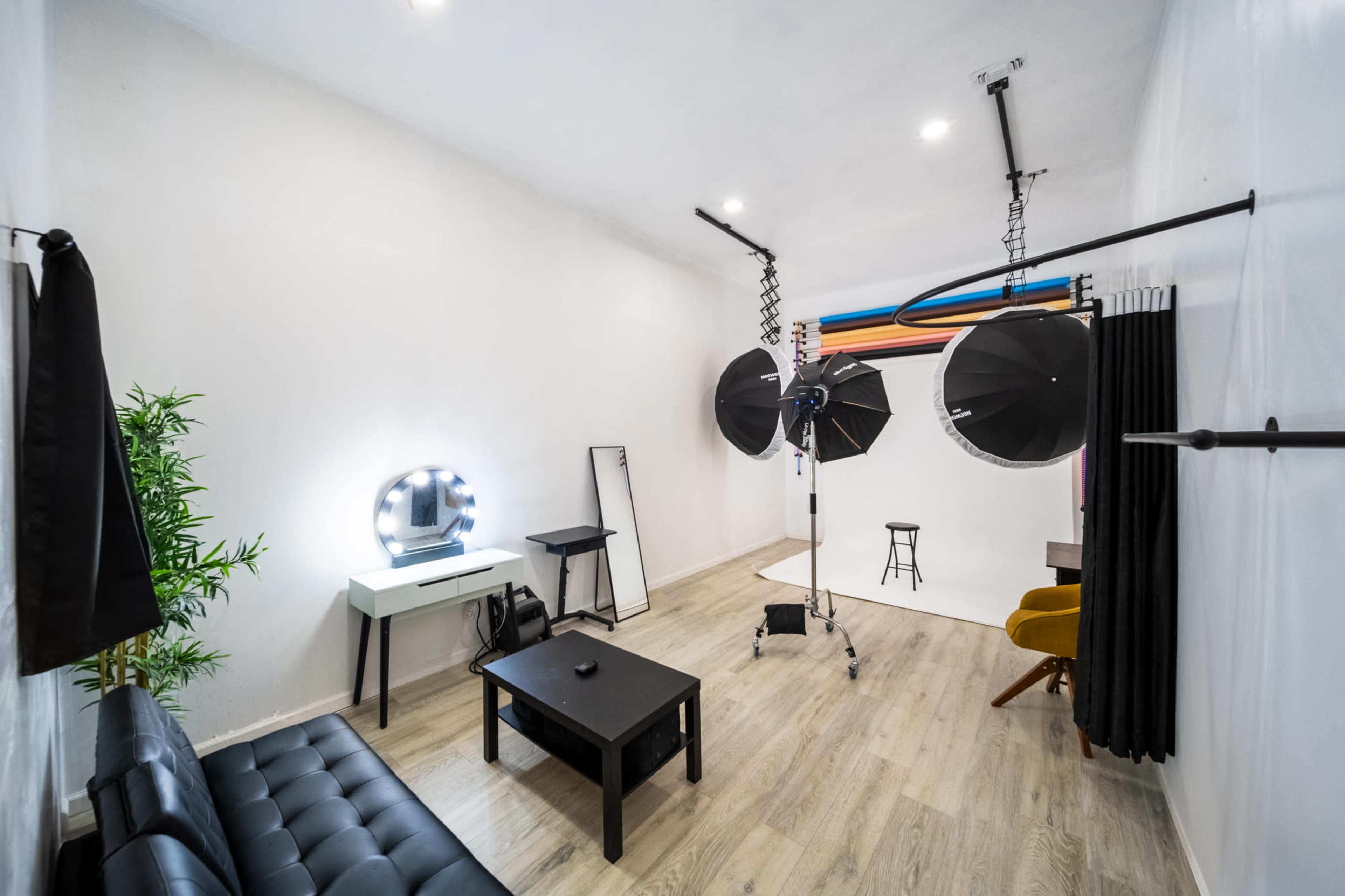 The image shows a photography studio with lighting equipment, a stool, a mirror, and a sofa arranged in a well-lit, minimalist space.