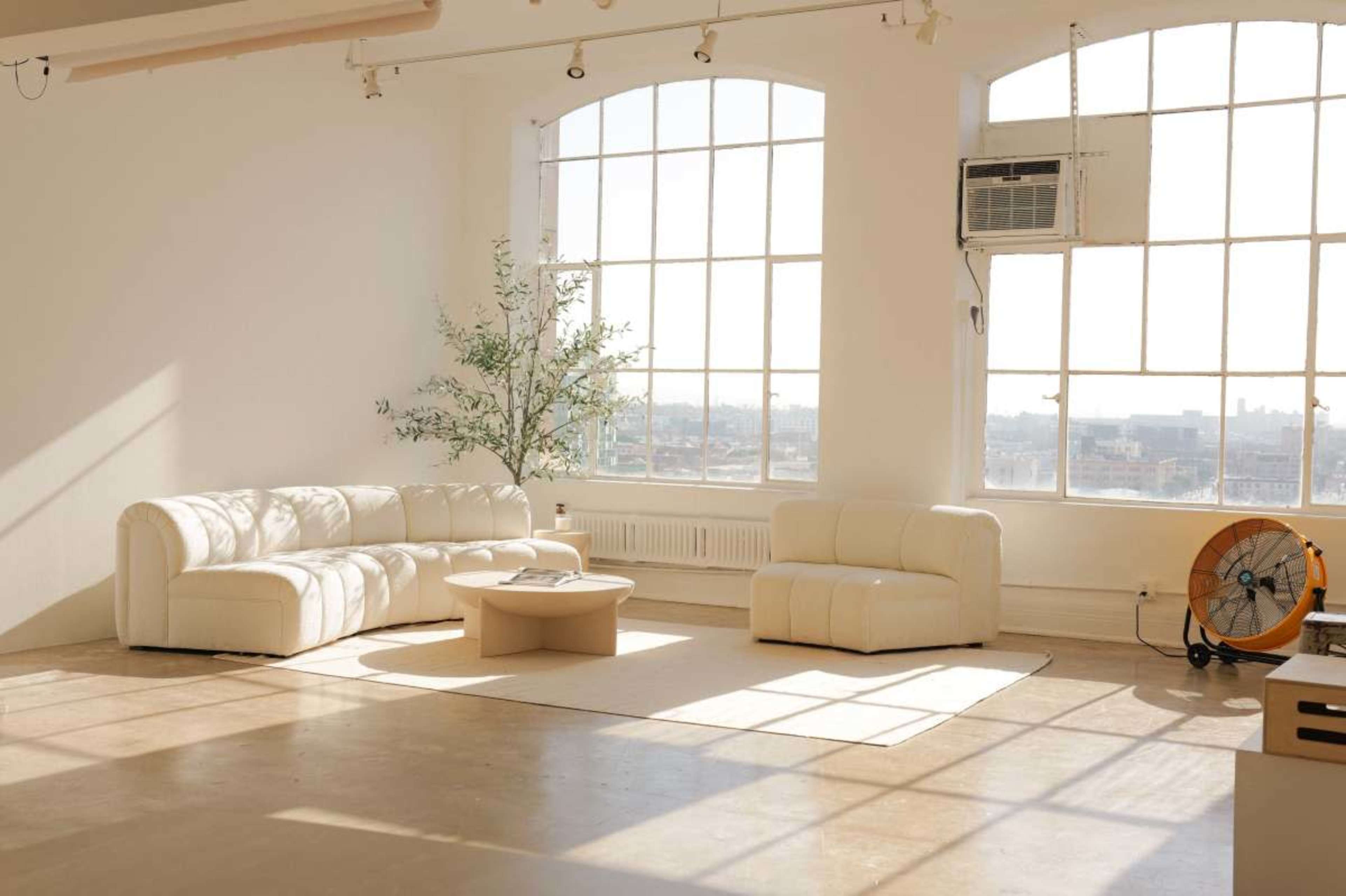 A spacious, airy room features a light-colored sofa and armchair, a round coffee table, and large windows that provide a view of the city.
