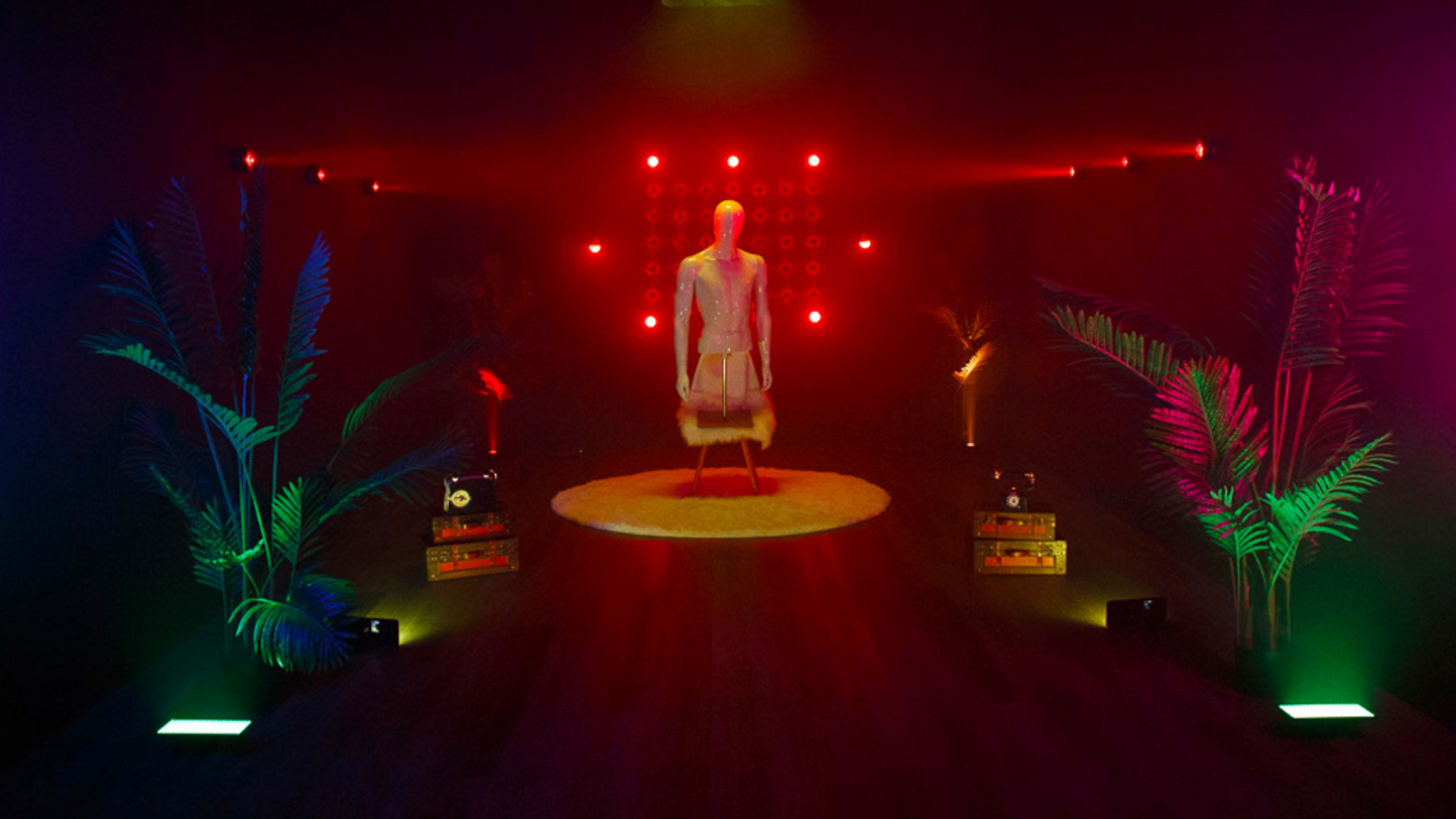 A mannequin stands on a circular rug in a dimly lit room, surrounded by colorful lights, palm plants, and speaker boxes.