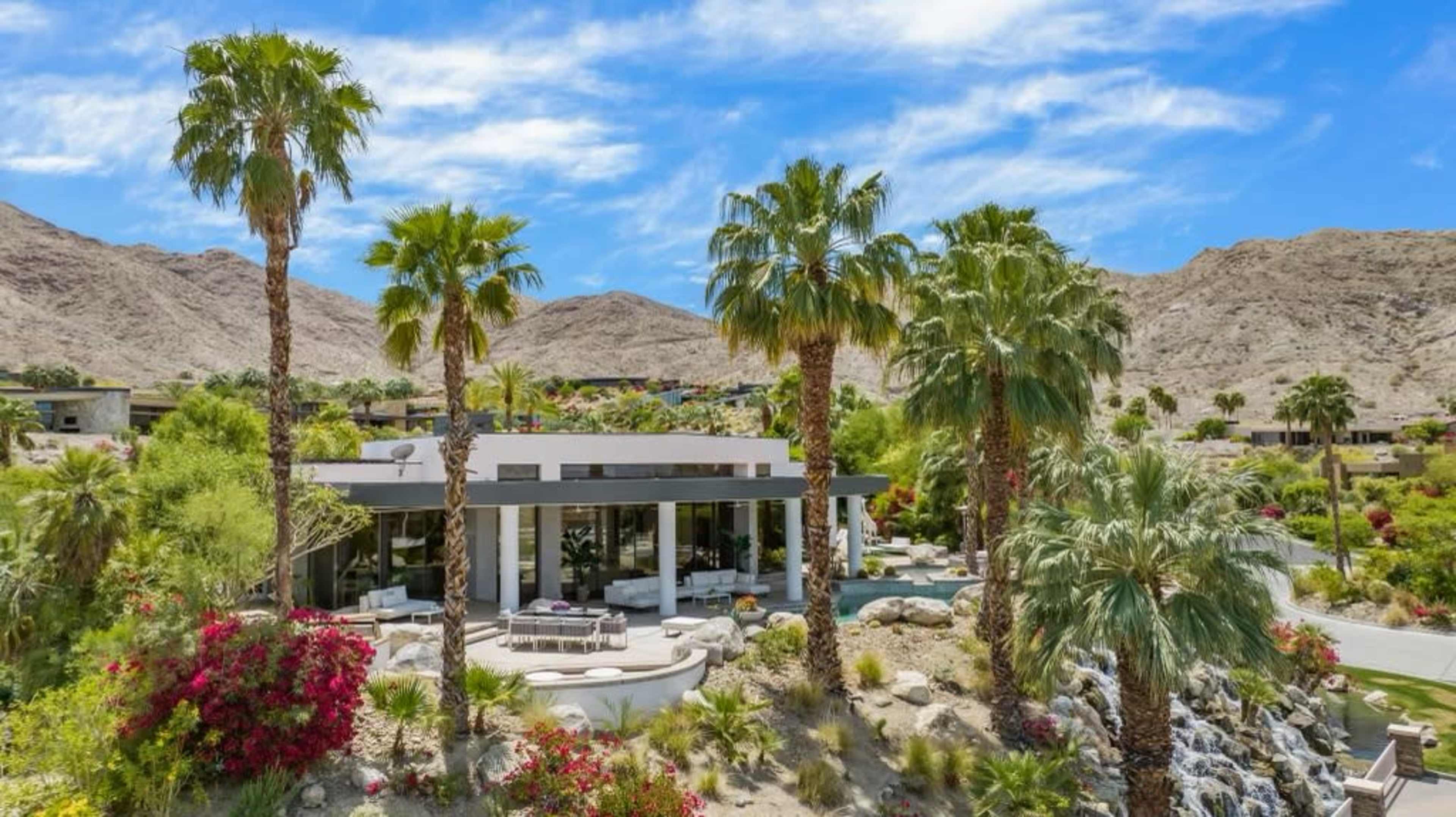 The image shows a modern house surrounded by palm trees and lush vegetation, set against a backdrop of mountains.