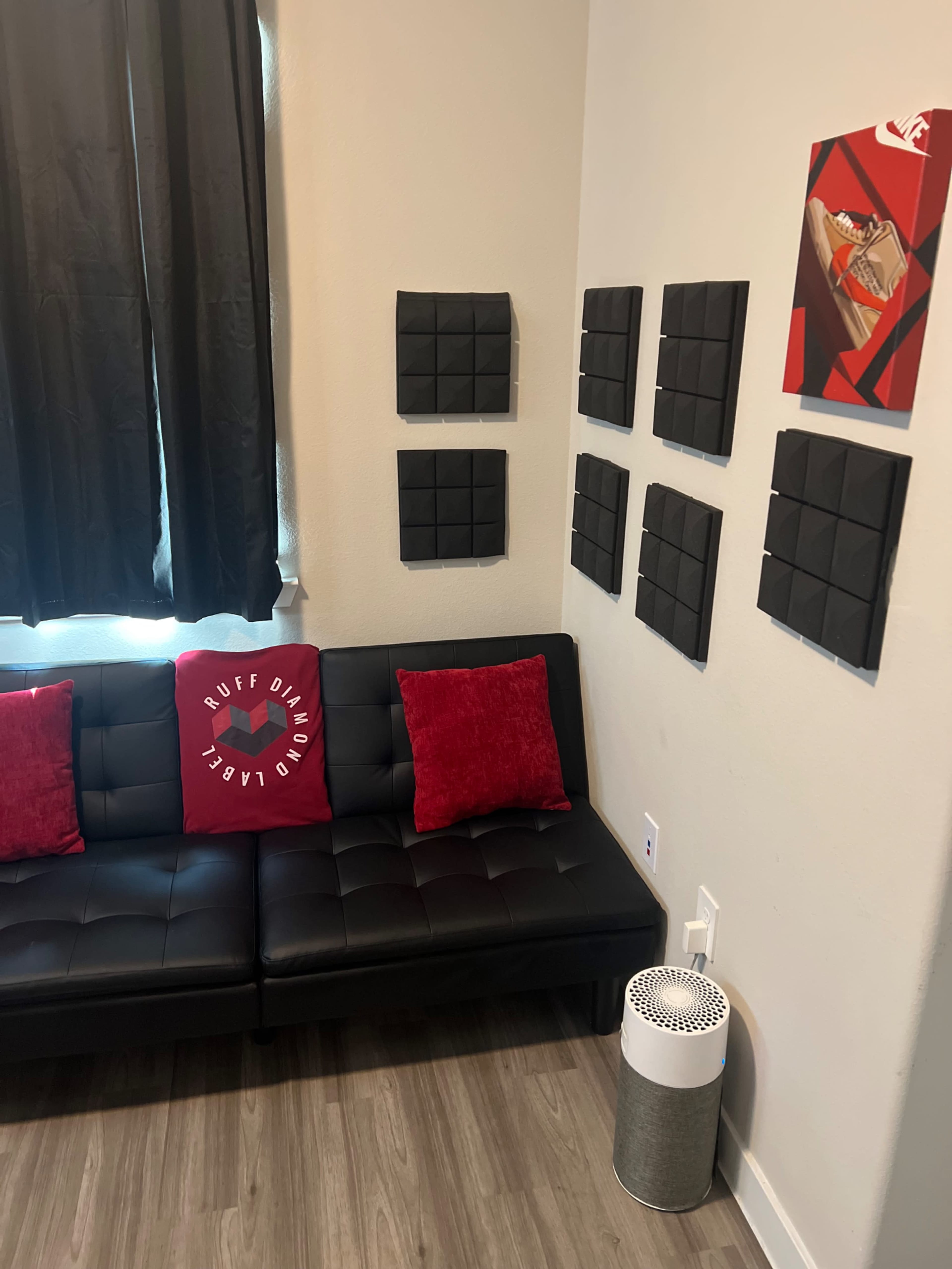 A black couch with red pillows sits against a wall adorned with black sound panels, beside a white air purifier and a piece of artwork.