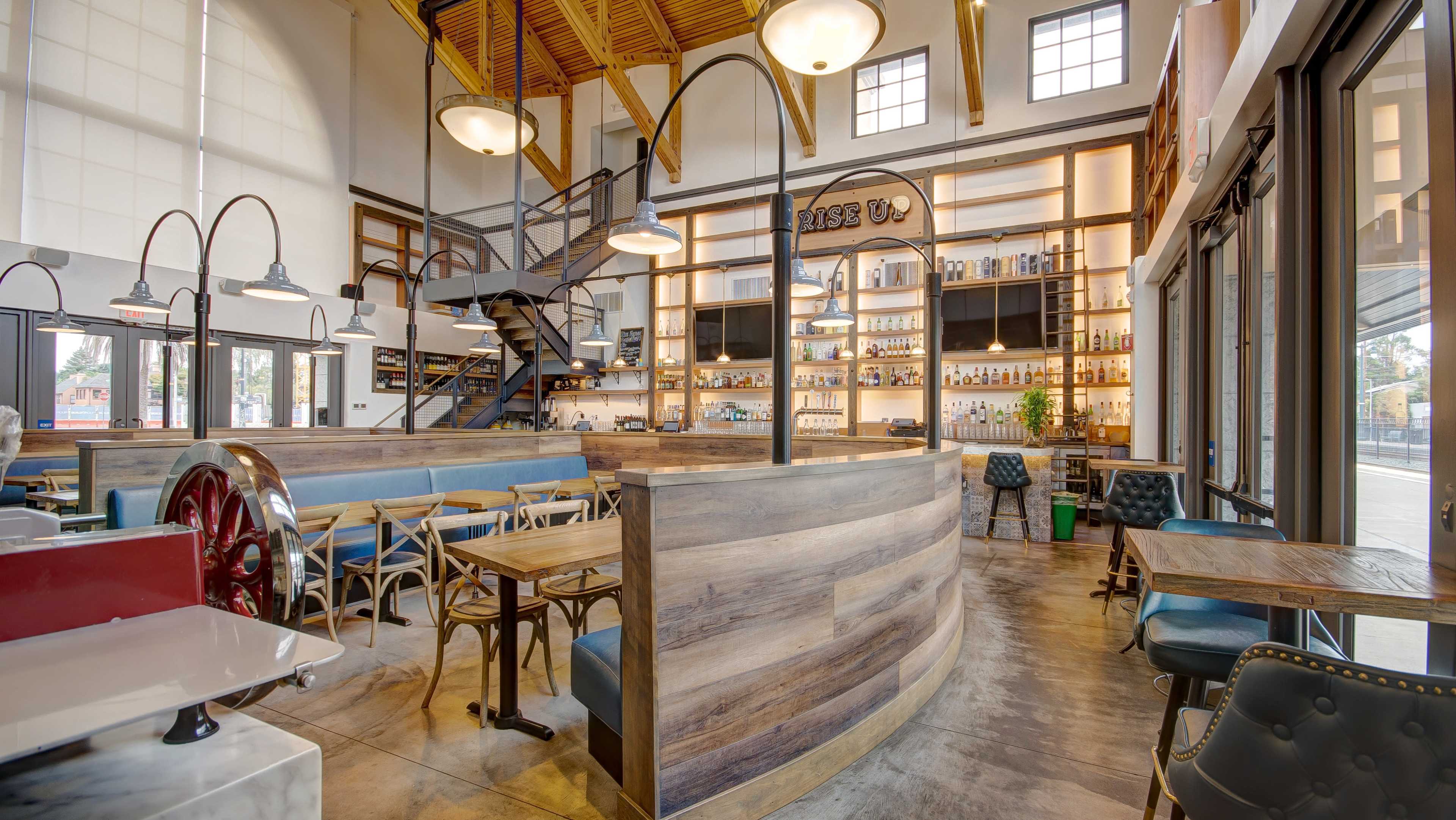 A modern restaurant interior featuring wooden furnishings, a curved bar area, and a staircase leading to a second floor, with shelves displaying an array of liquor bottles.