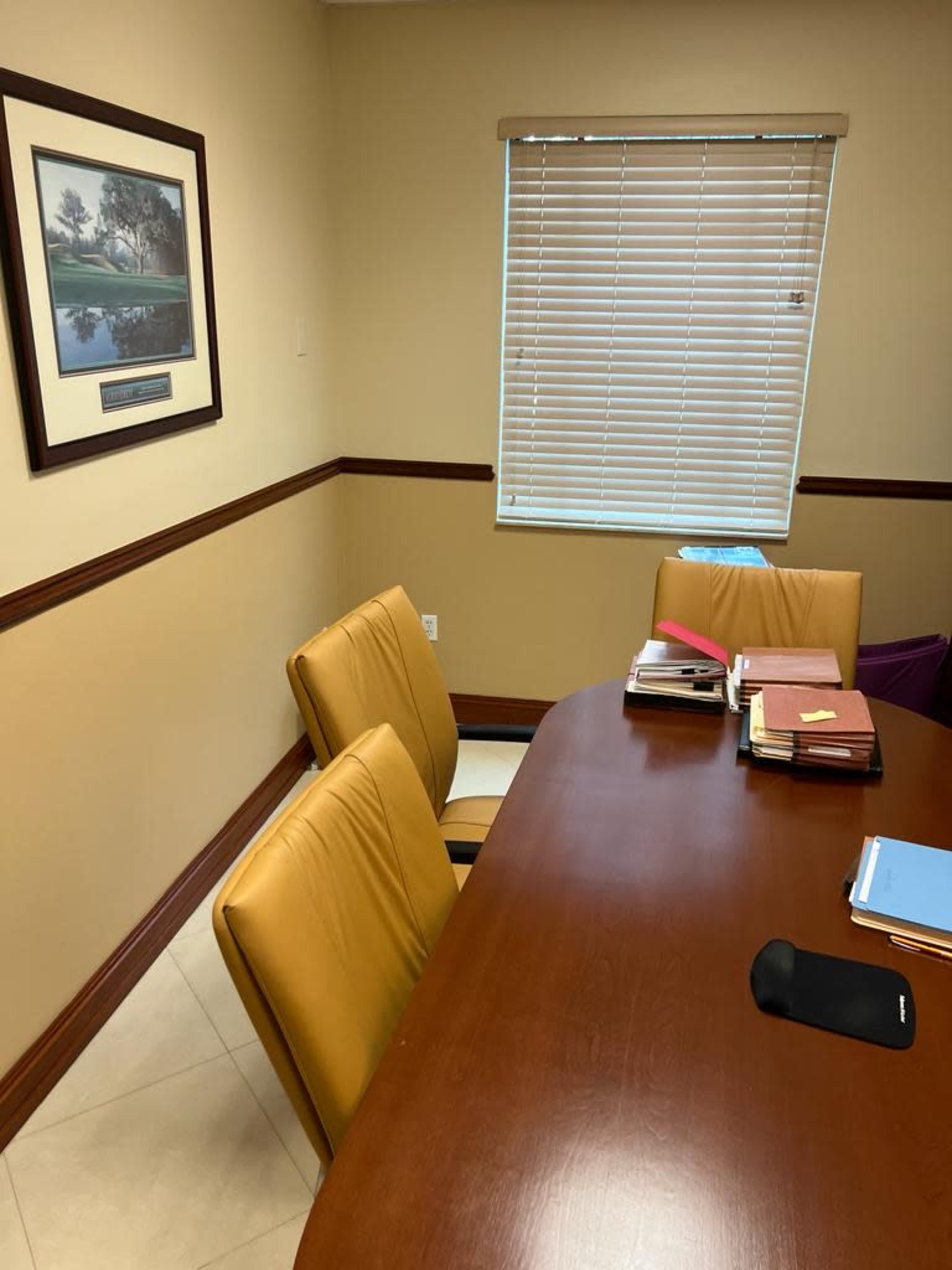 A corner of an office with a brown wooden table, two yellow chairs, a stack of assorted files, and a window with blinds partially open.