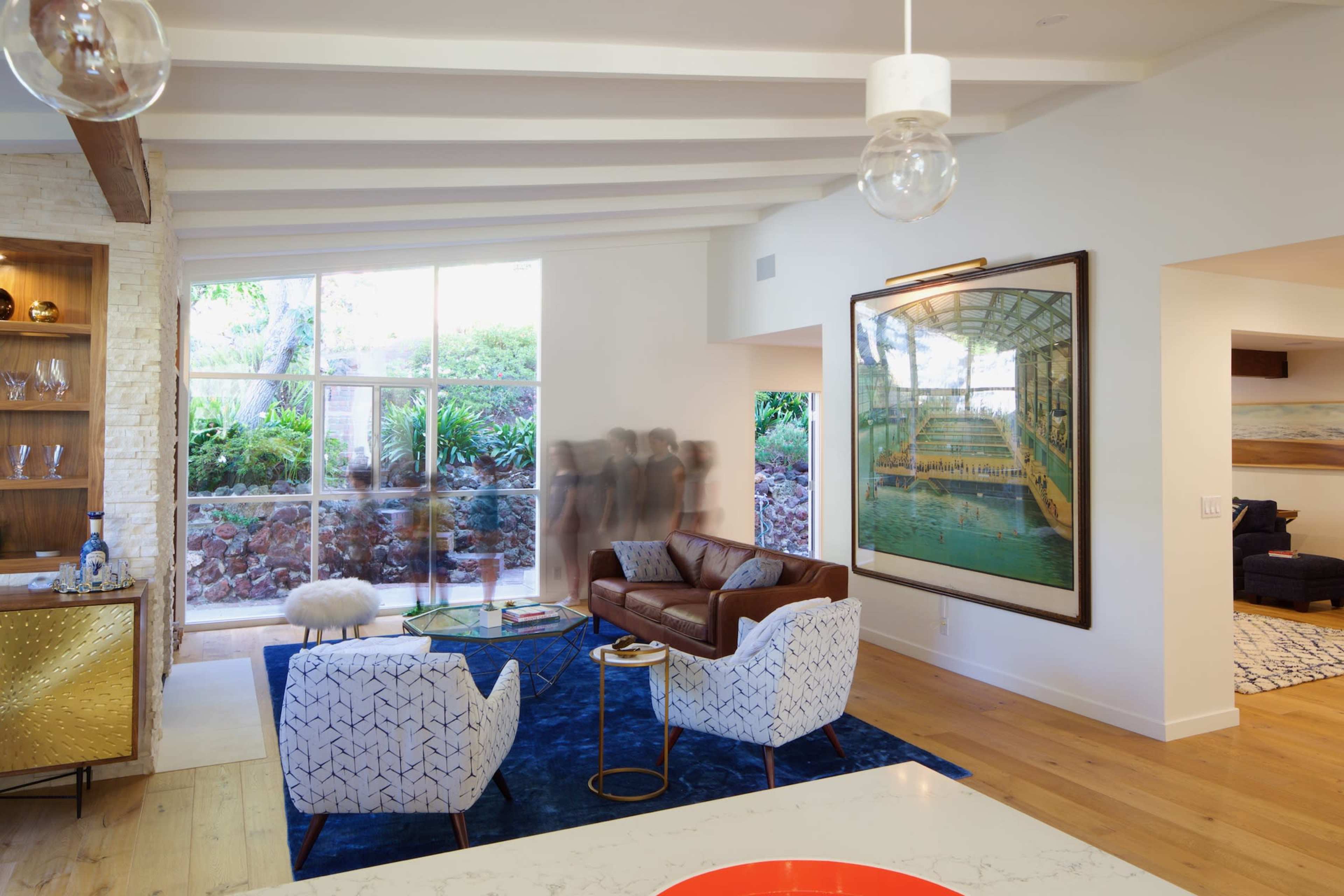 A modern living room with large windows, a brown leather sofa, two patterned chairs, and a large framed painting on the wall.