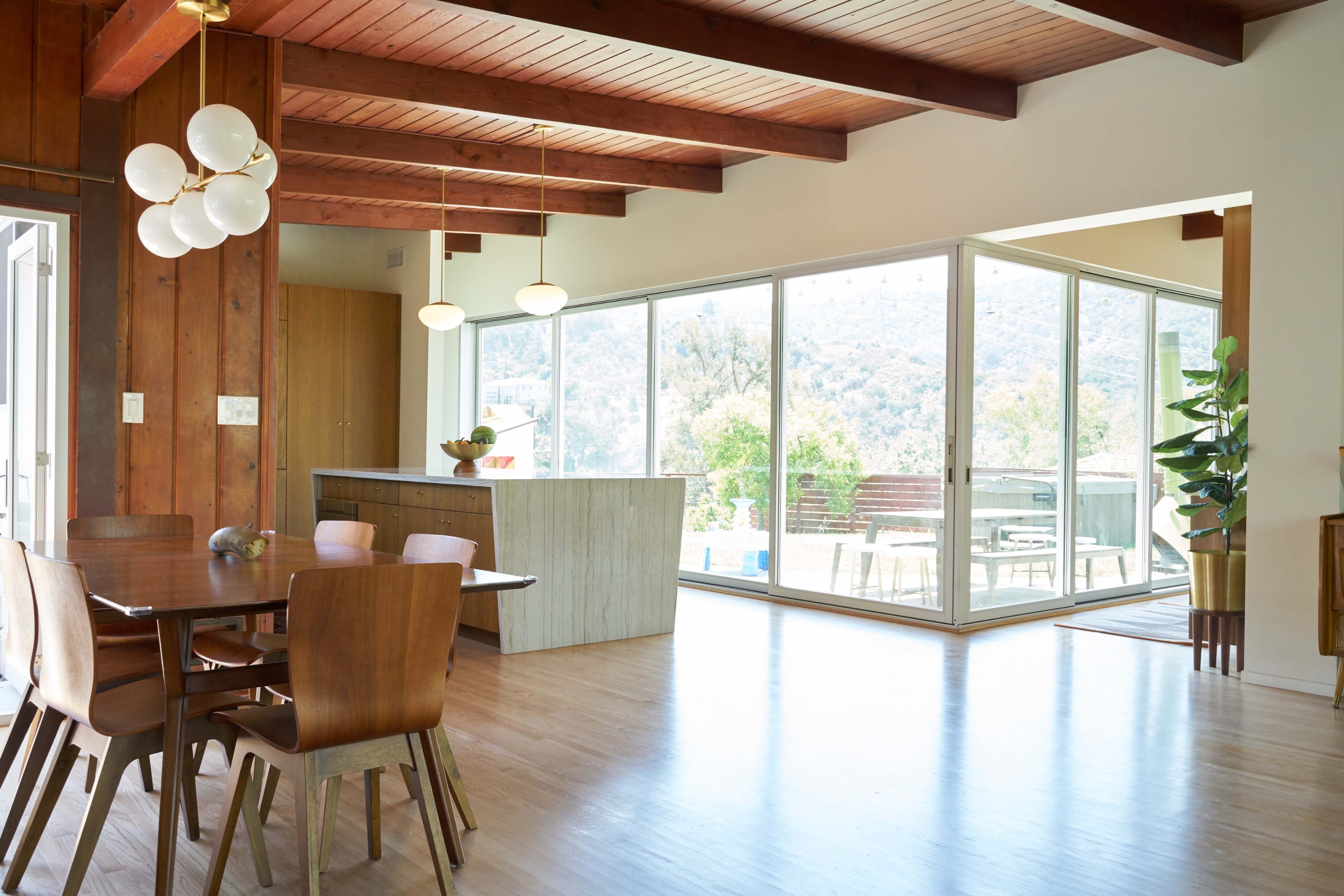 A modern dining area features a wooden table with chairs, a large glass window opening to an outdoor space, and minimalist decor including a stone kitchen island.