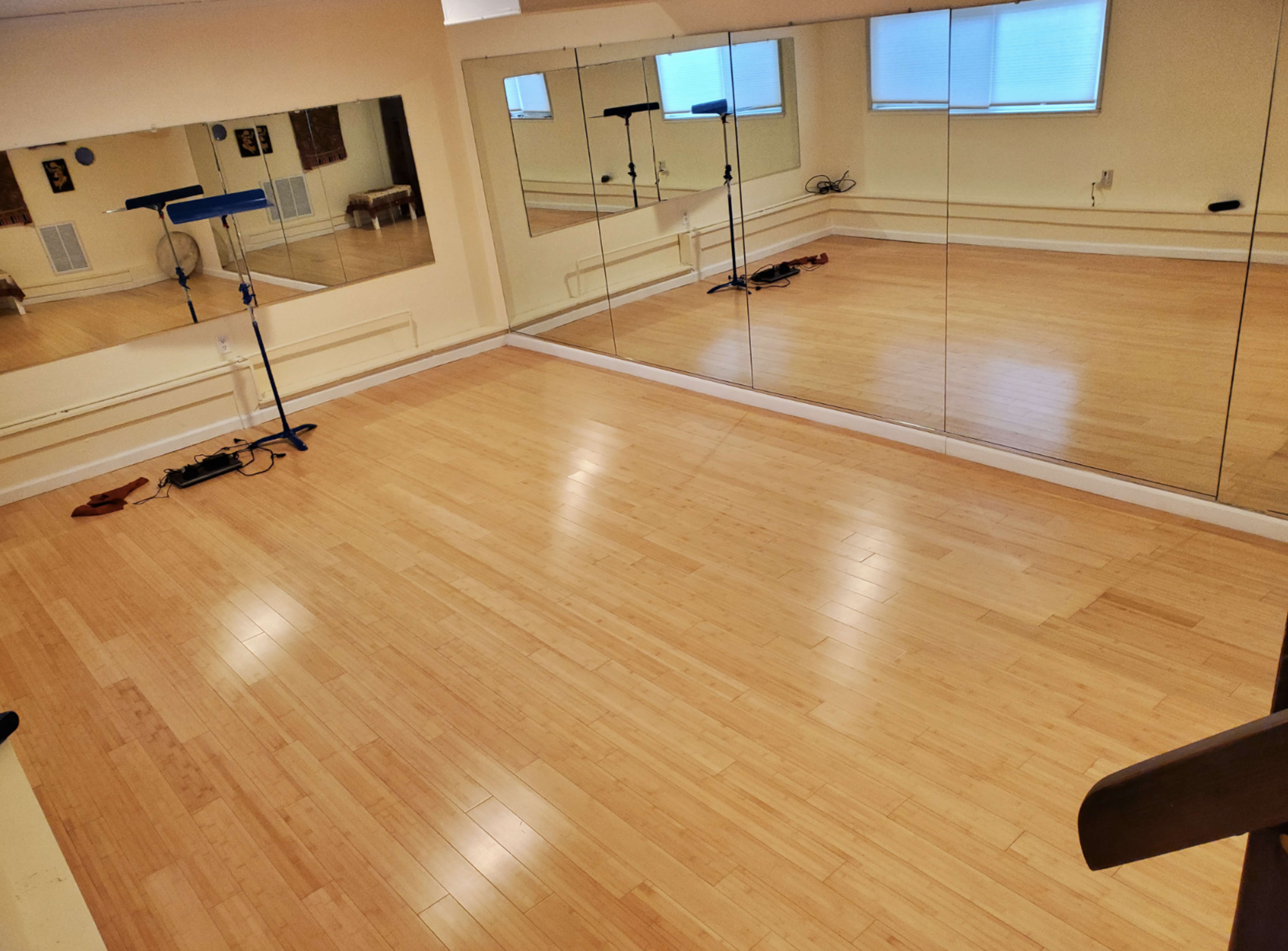The image shows a spacious room with wooden flooring and large mirrors along one wall, illuminated by natural light from a window.