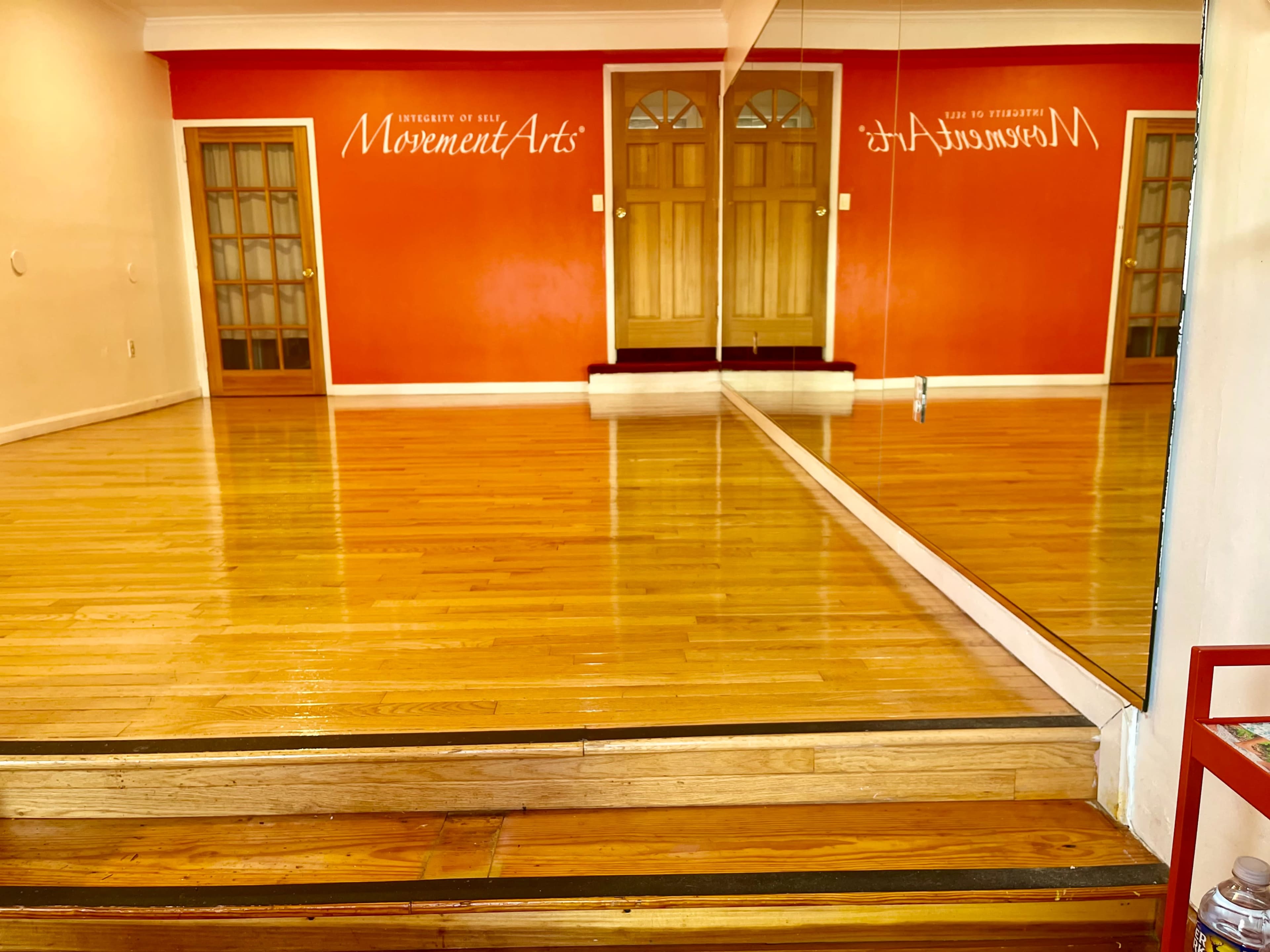The image shows a dance studio with a polished wooden floor, a red wall featuring the text "MovementArts," and a large mirror reflecting the space.