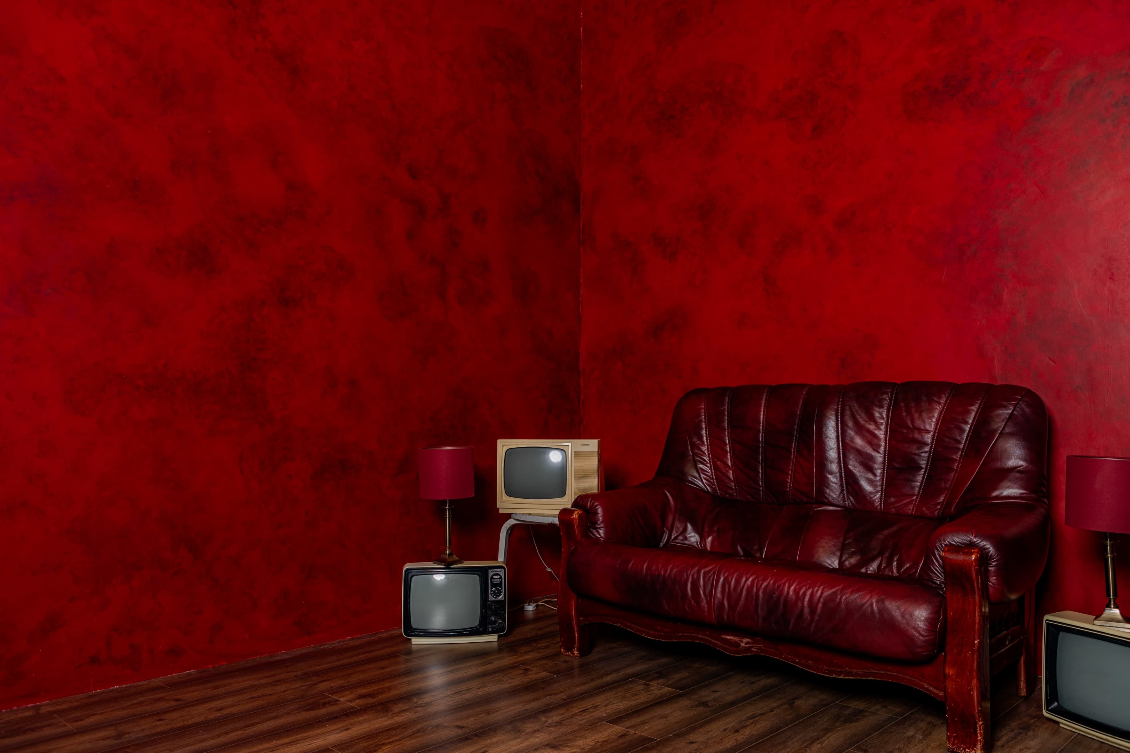 The image shows a red-walled room featuring a leather couch, two small lamps, and two vintage televisions.