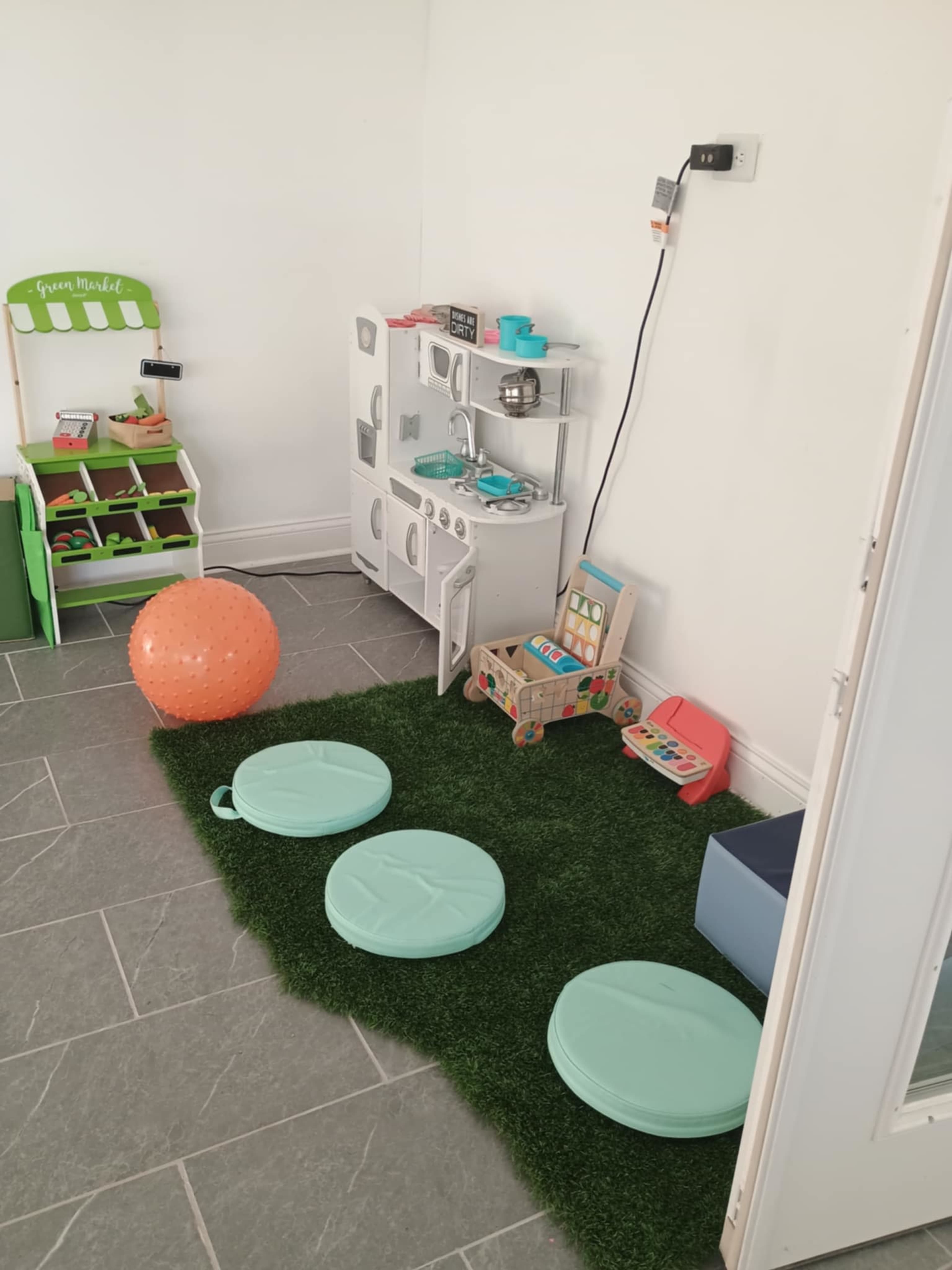 The image shows a play area featuring a small kitchen set, colorful toys, a large orange ball, and green circular seating on faux grass tiles.