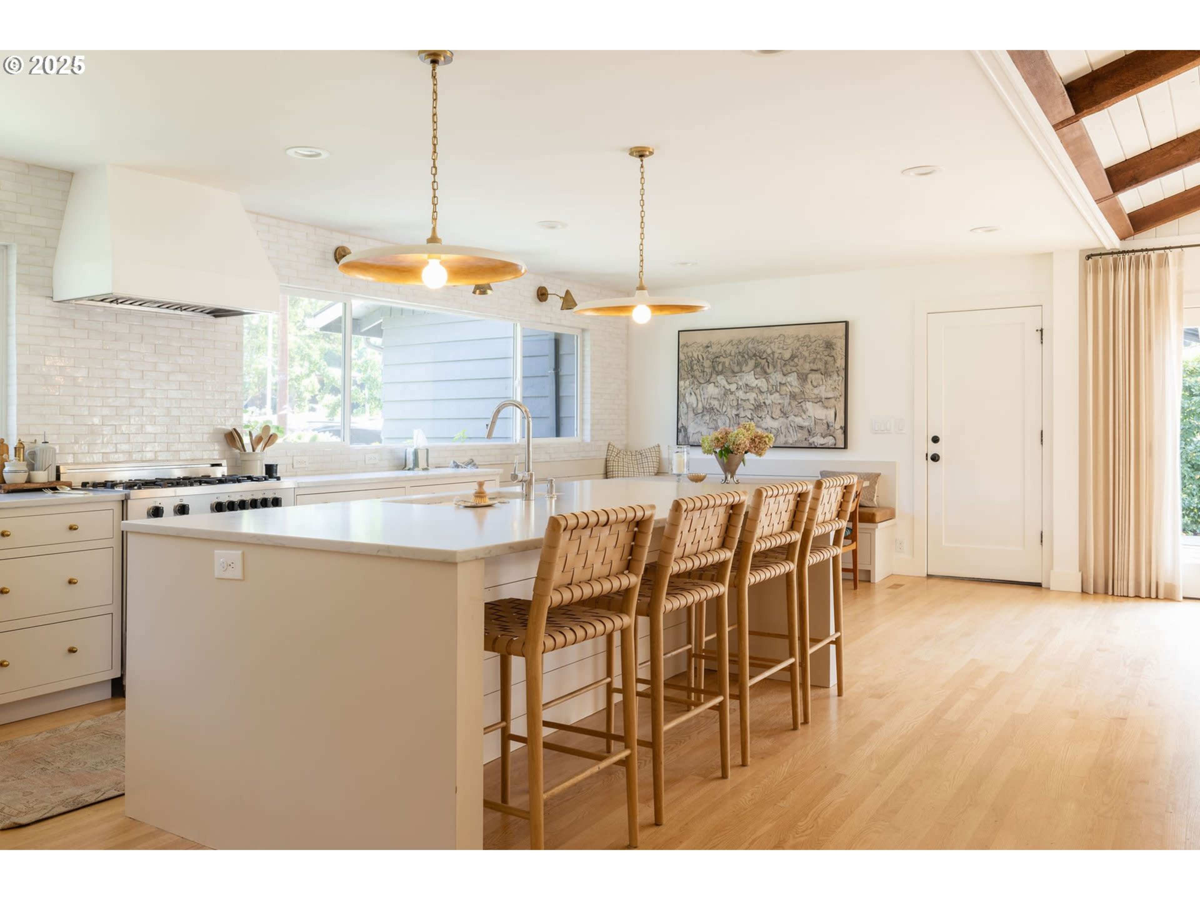 The image shows a modern kitchen featuring a large island with bar seating, pendant lights, and a window allowing natural light.