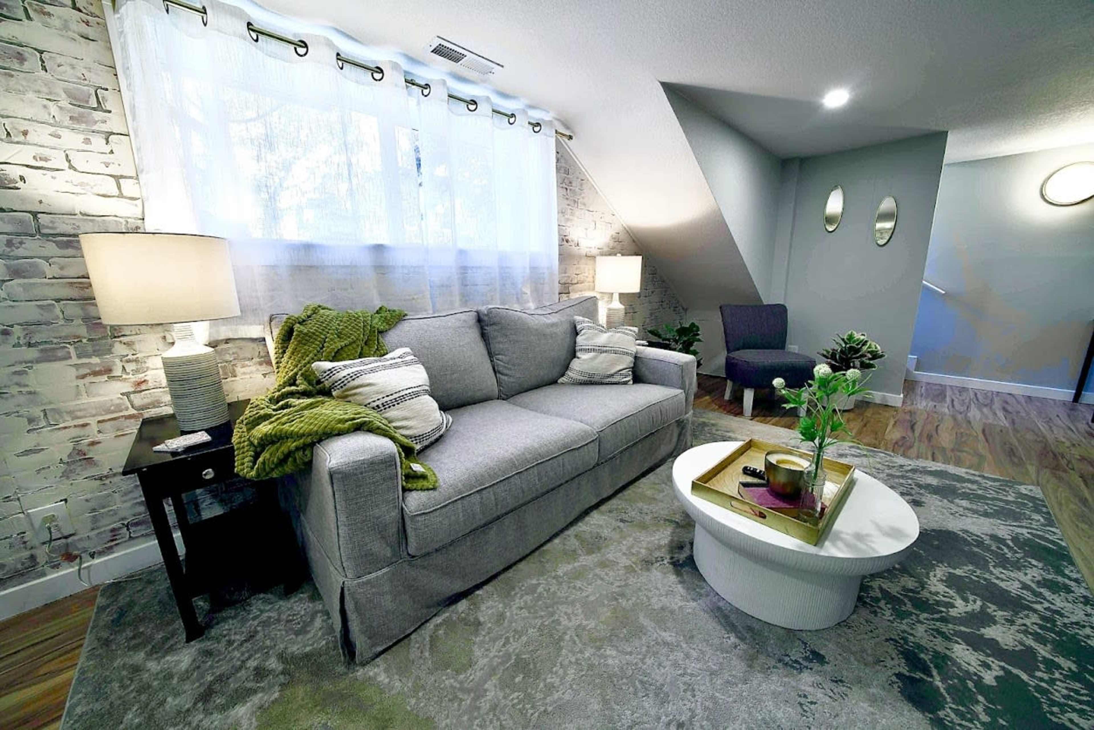 A cozy living room setup with a gray sofa, a round coffee table, and a small armchair against a backdrop of brick-patterned walls and large windows.
