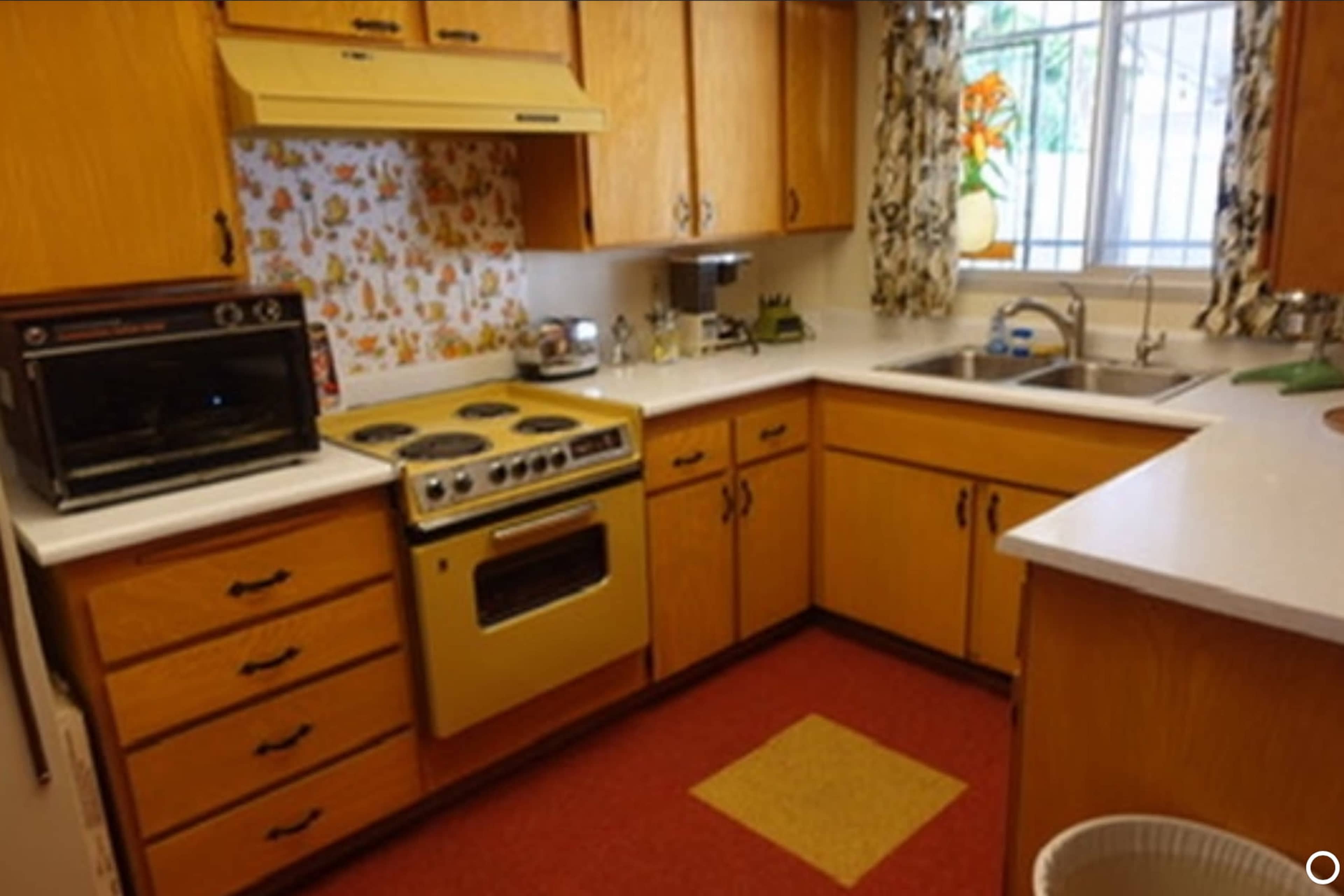 The image shows a compact kitchen with wooden cabinets, a yellow stove, and a sink under a window, featuring a patterned backsplash and a mix of countertop appliances.