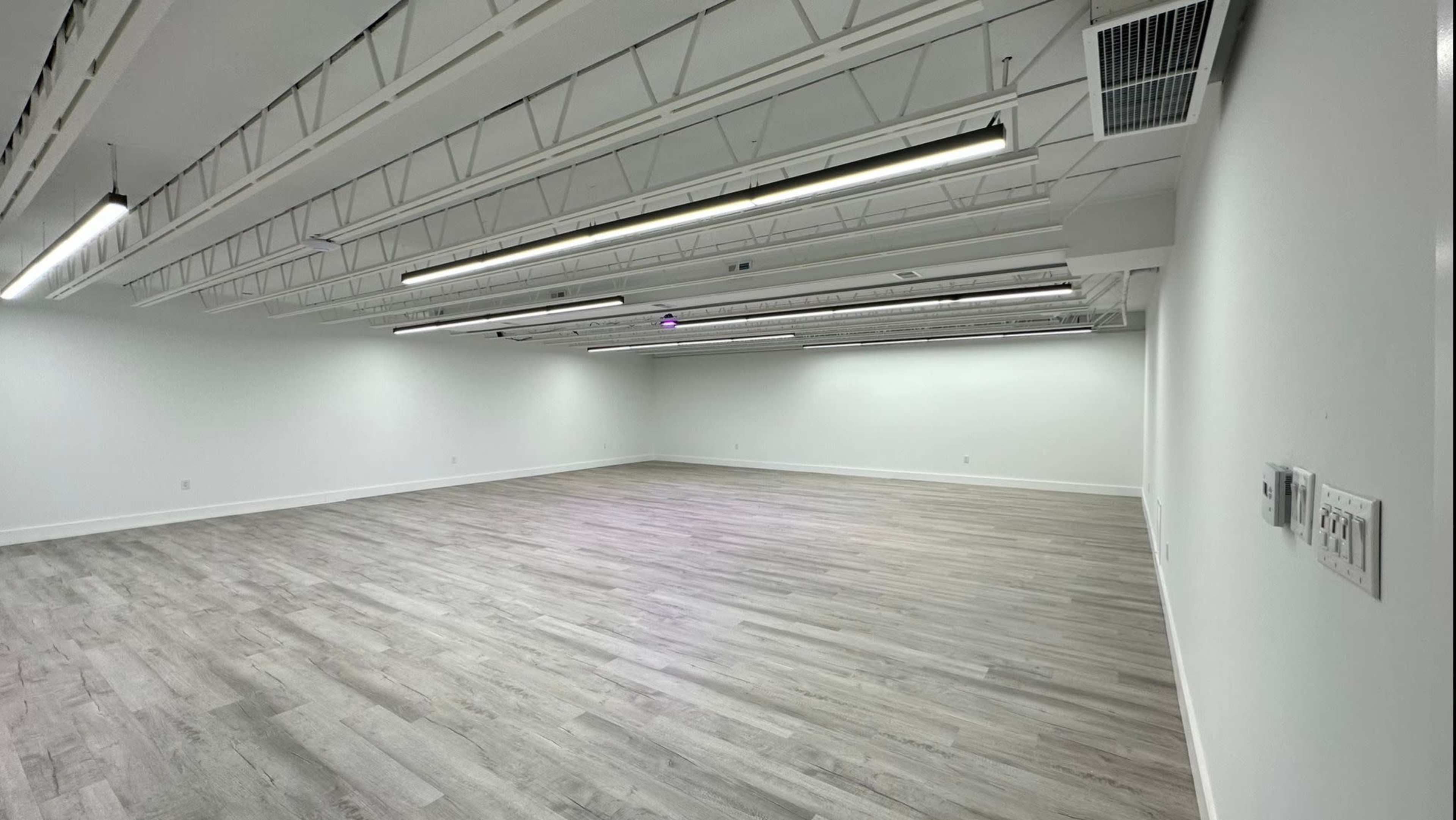 An empty room with white walls, a light wooden floor, and overhead lighting fixtures.