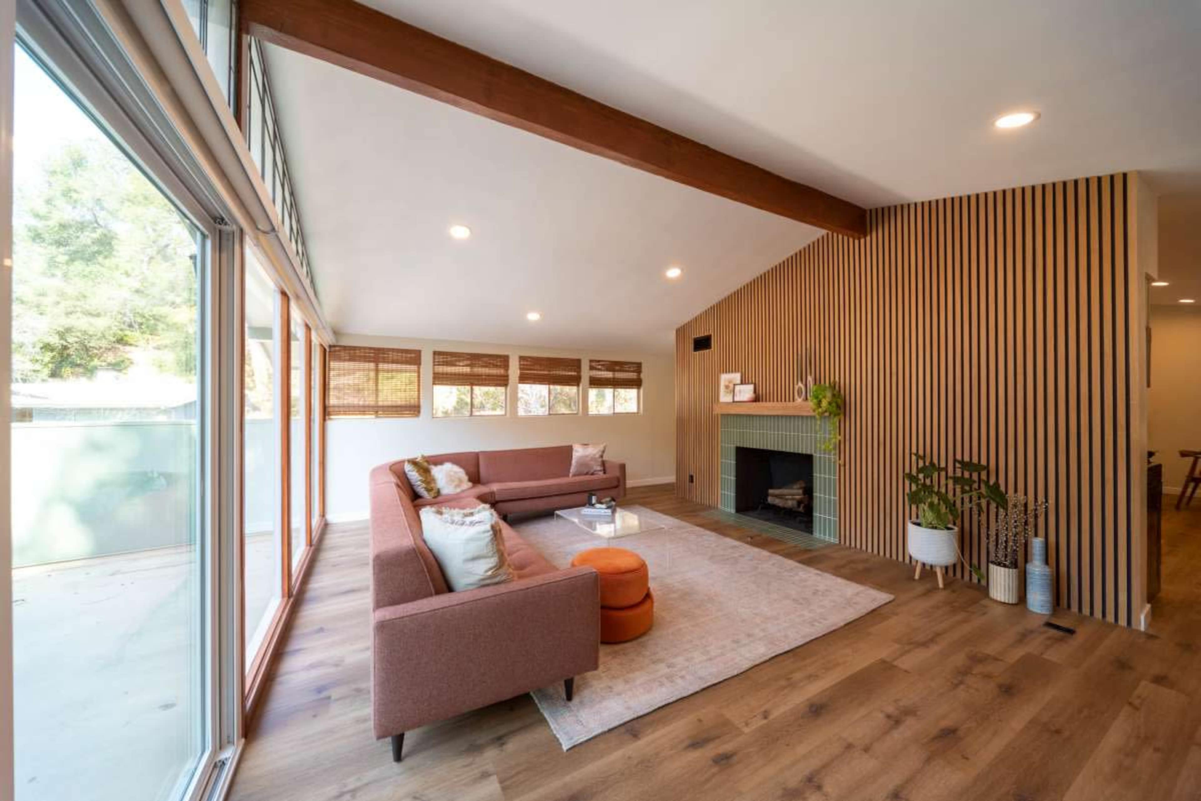 A modern living room features a brown sectional sofa, a round accent table, a fireplace, and wood-paneled walls with large windows allowing natural light.