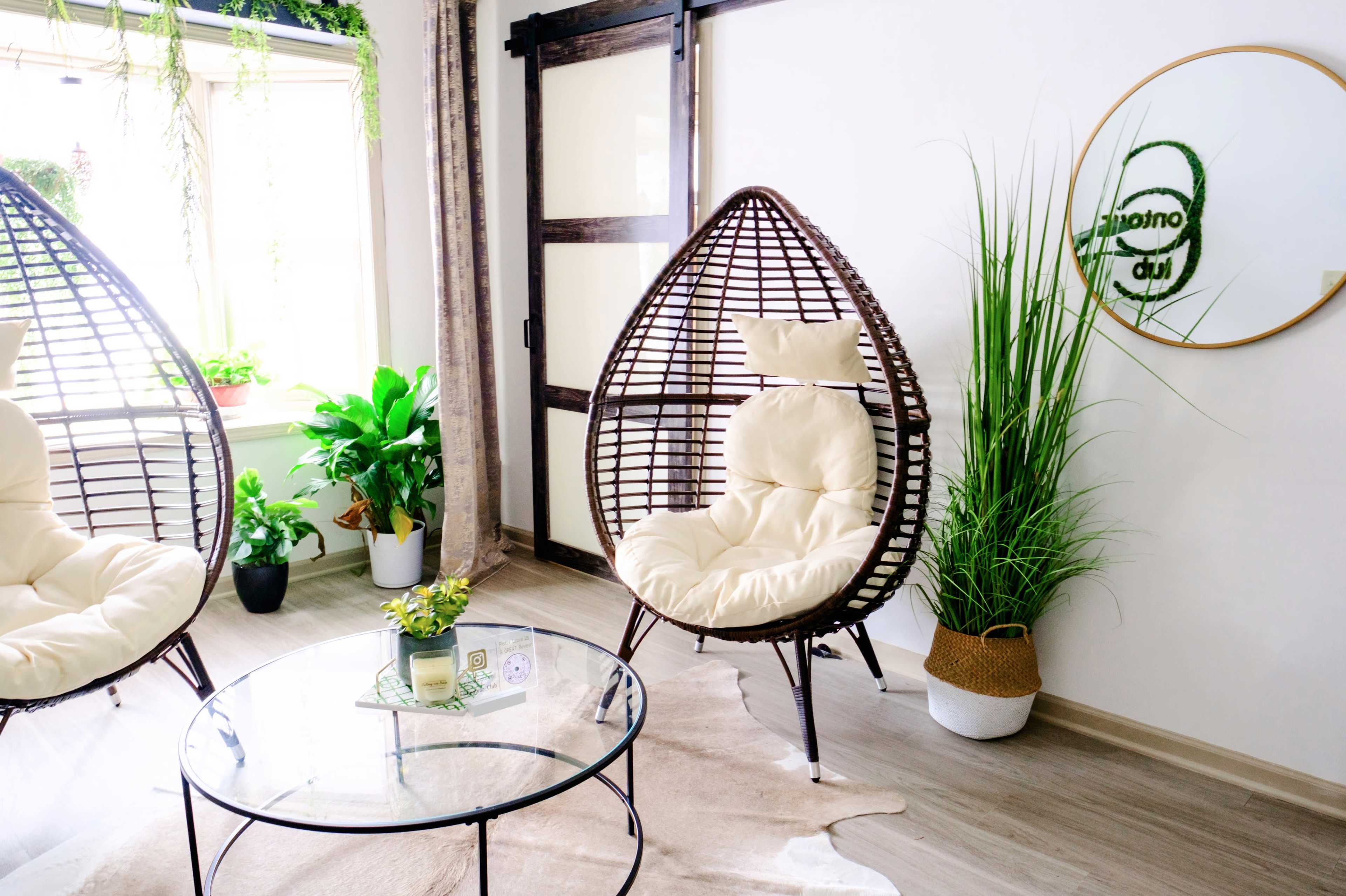 A bright, modern living room featuring two wicker egg chairs with light cushions, a glass coffee table, and indoor plants beside a large window.