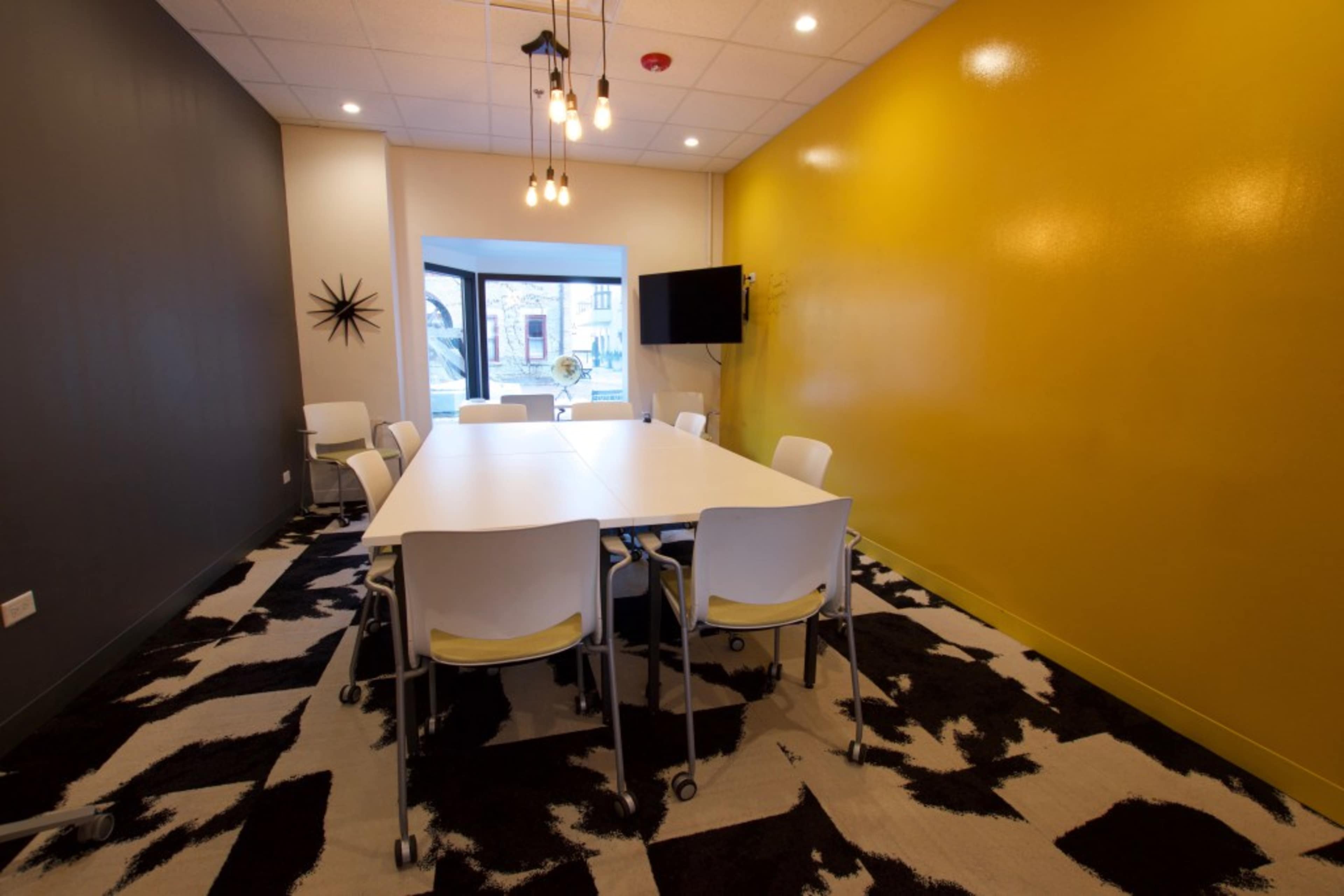 A modern meeting room features a large white table surrounded by eight rolling chairs, with a yellow accent wall and a window providing natural light.