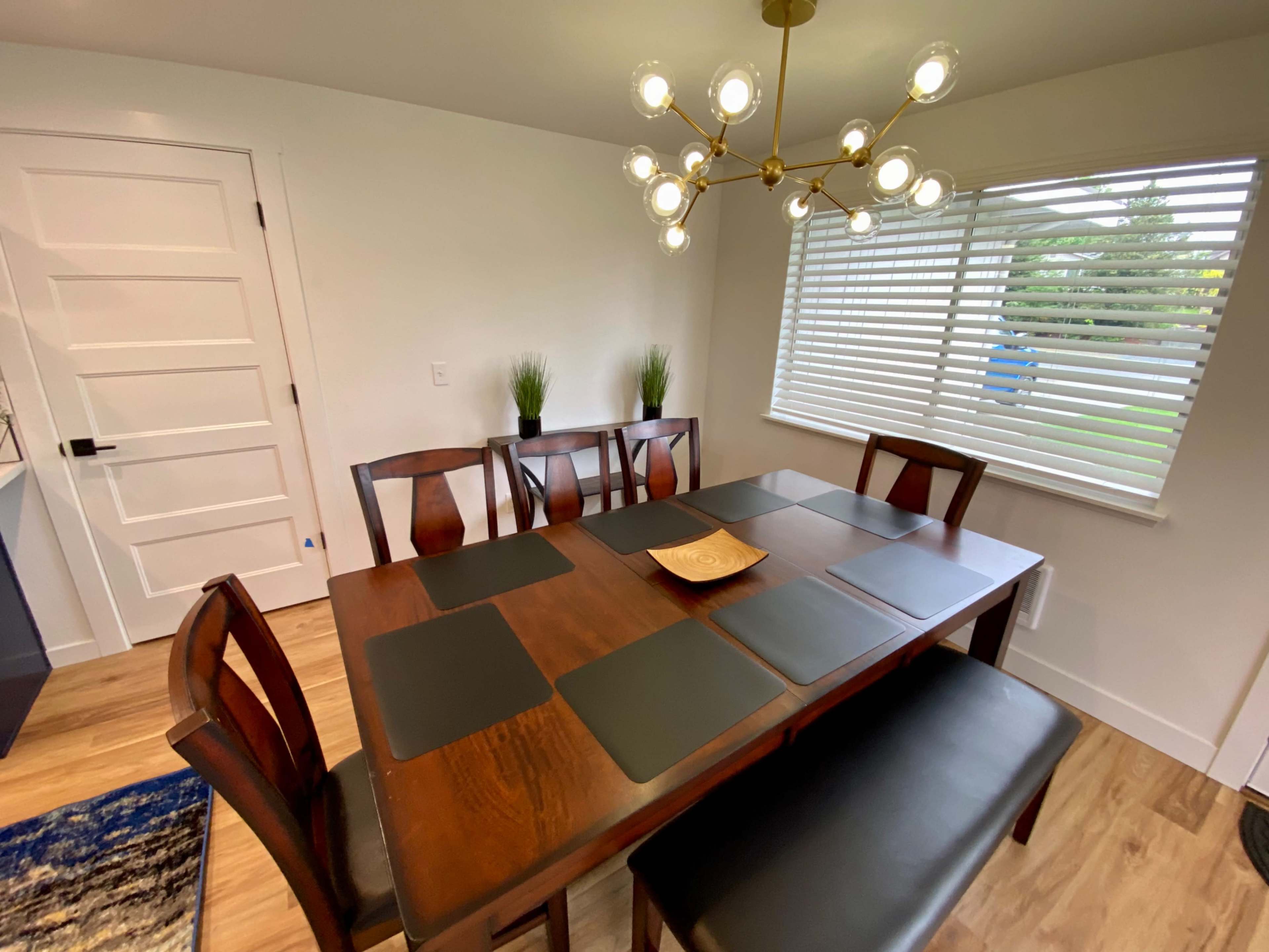 A modern dining room featuring a wooden table with chairs, a decorative light fixture, and a window with blinds.