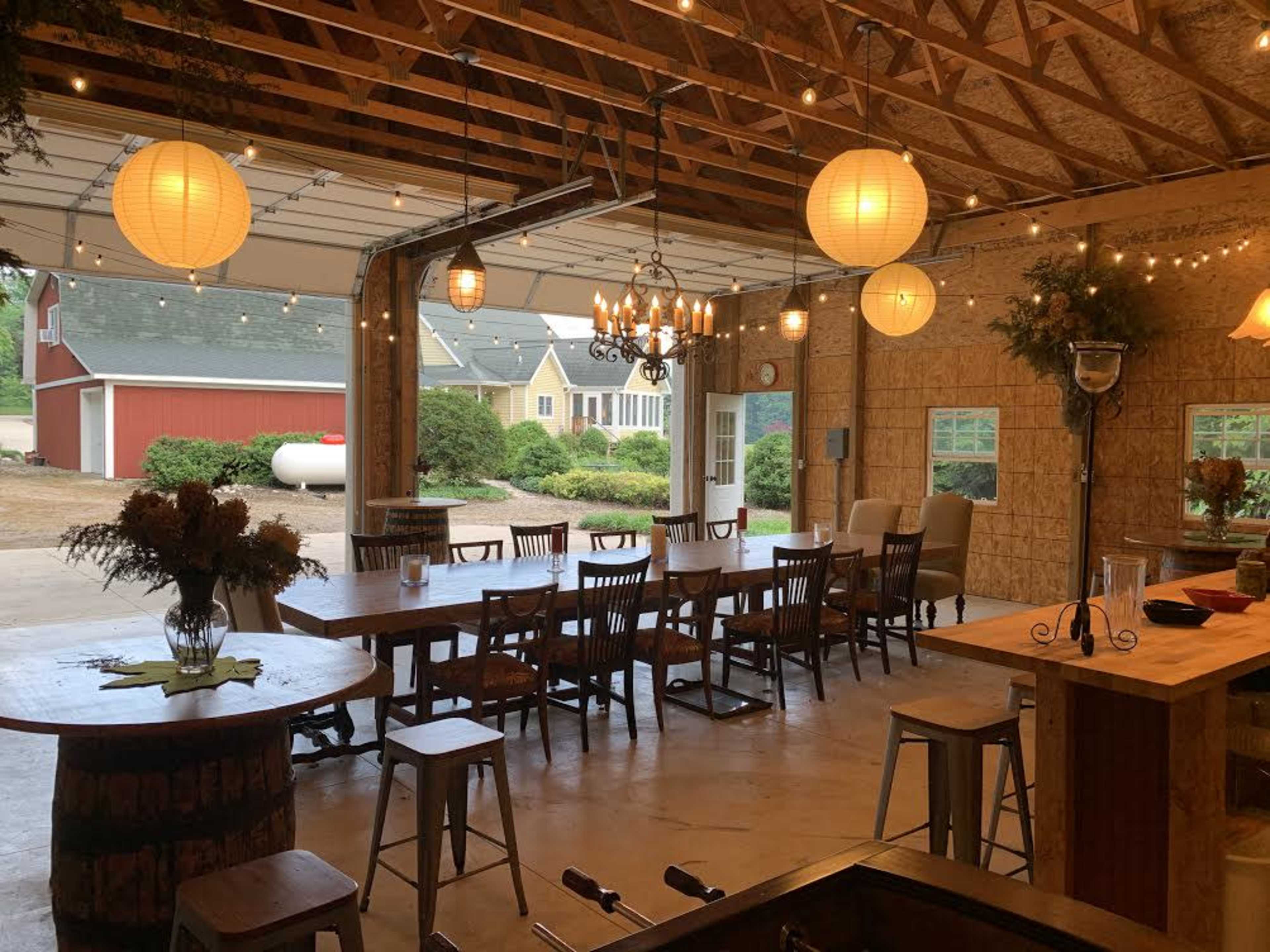 A spacious gathering area with a long wooden table, surrounded by chairs, illuminated by string lights and paper lanterns, featuring an open garage door that reveals an exterior view of a garden and nearby buildings.