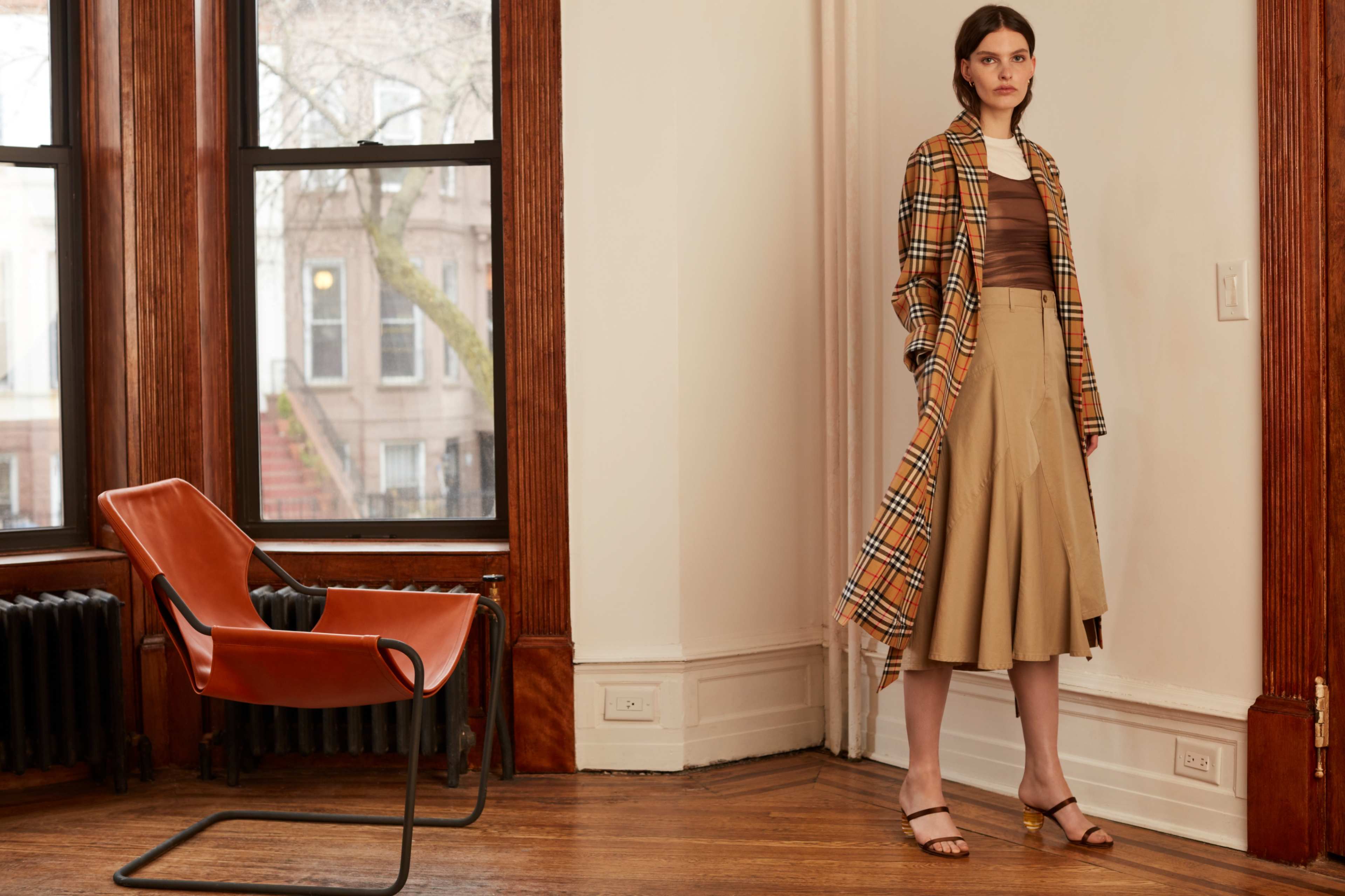 A woman stands in a room with large windows, wearing a plaid coat over a brown top and a beige skirt, next to a modern orange chair.