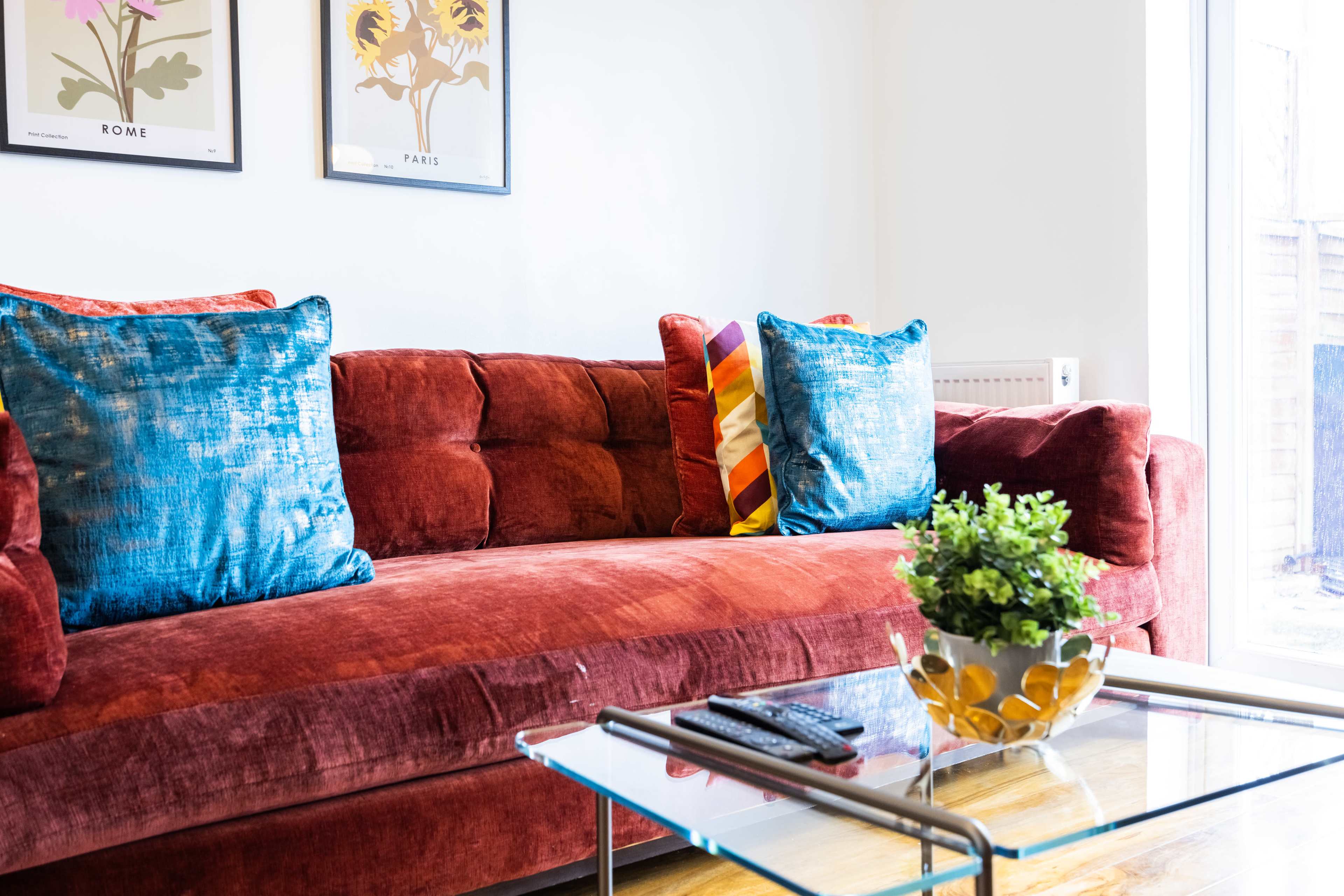 A plush red sofa with decorative blue and colorful cushions is positioned next to a glass coffee table with a small potted plant.