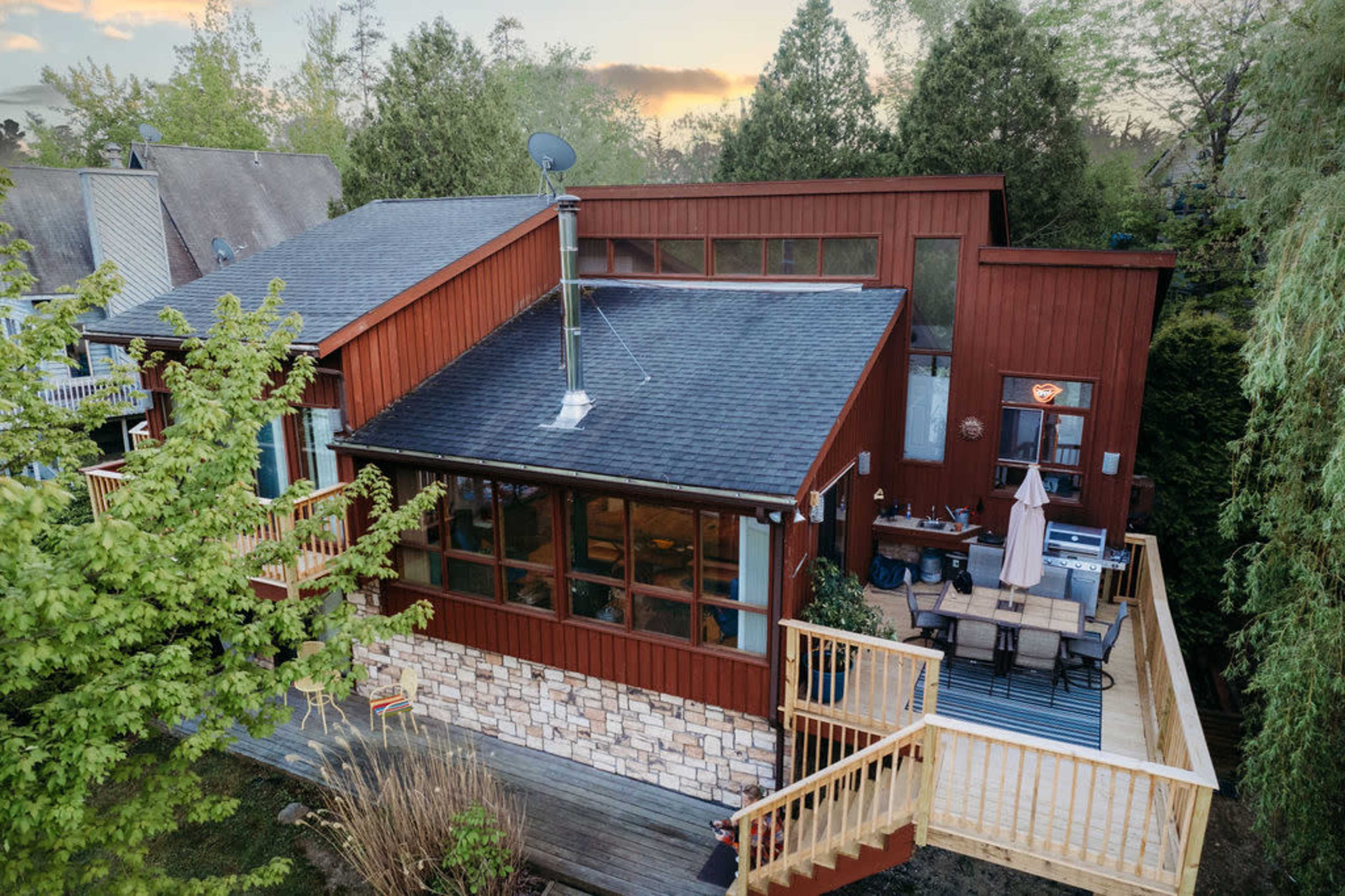 A modern house with a red wooden exterior features a deck, large windows, and a chimney, surrounded by trees and greenery.